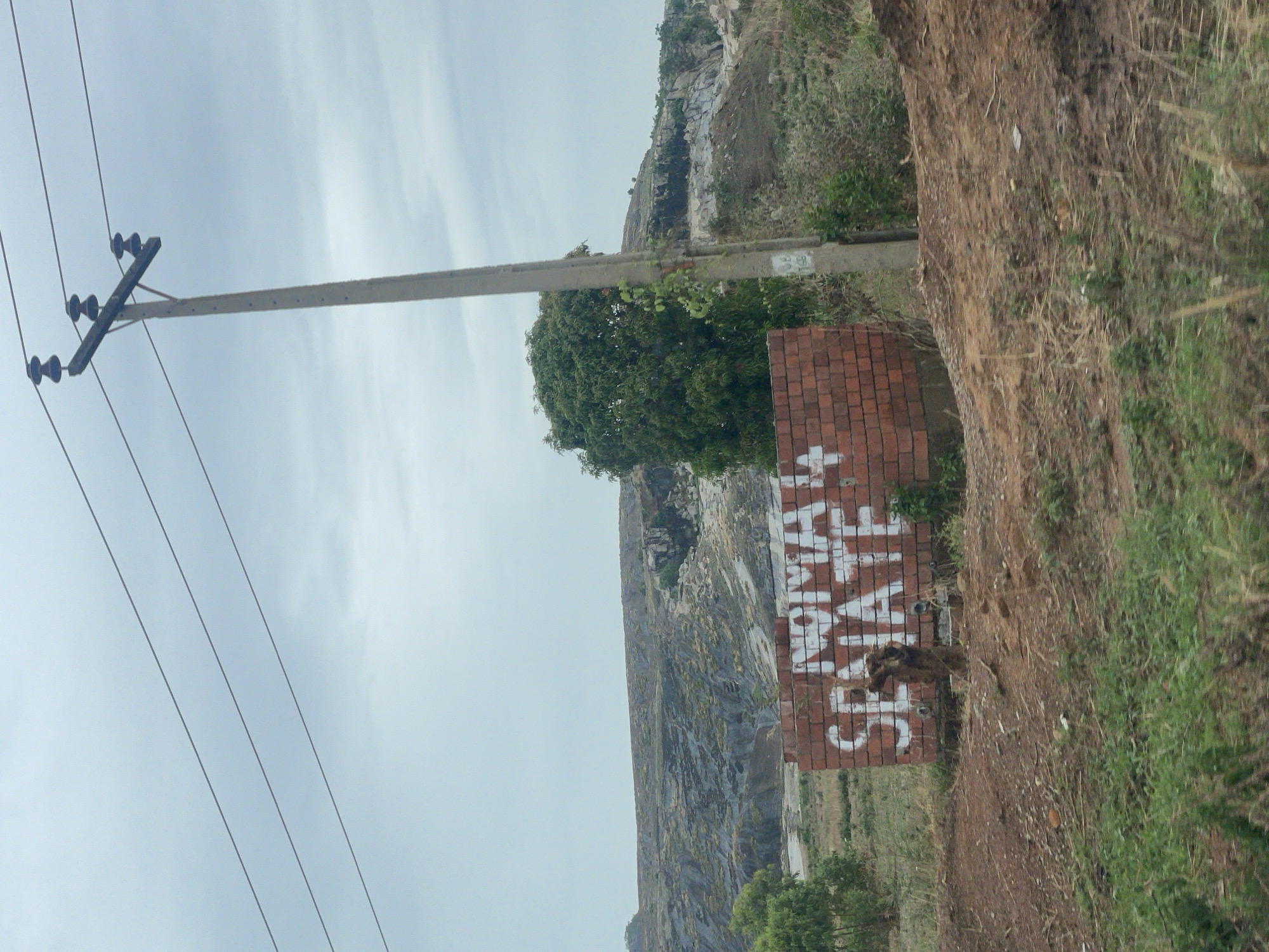 A brick wall stands beside a dirt road at the base of a rocky slope, with graffiti "Skate Novam" painted on the wall. A utility pole is positioned on the left side of the image. The scene is located near the quarry in Kujama, Nigeria.