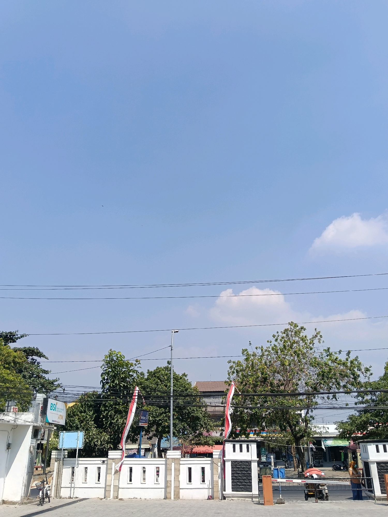 The image captures a street scene in Balun, Indonesia under a clear blue sky with scattered white clouds. In the foreground, a white wall with black and white decorative pillars lines the street. Indonesian flags, red and white, are prominently displayed, attached to poles near the entrance.
On the left side of the image, a white building with a sign that reads "BSI" and "ATM INDONESIA" is visible, indicating a bank. A bicycle is parked near this building. Behind the wall, trees with lush green foliage are abundant, with some buildings visible through the branches. Power lines crisscross the sky above the trees.
Further into the scene, there appears to be a barrier or gate across the road, with a motorcycle parked near it. Two individuals are present: one person, possibly a woman in a blue shirt and headscarf, stands near the barrier on the right side, and another person in a brown headscarf and blue shirt is partially visible near the motorcycle. The street surface is paved with gray interlocking blocks. The overall impression is a daytime view of a commercial or public area in an Indonesian town.