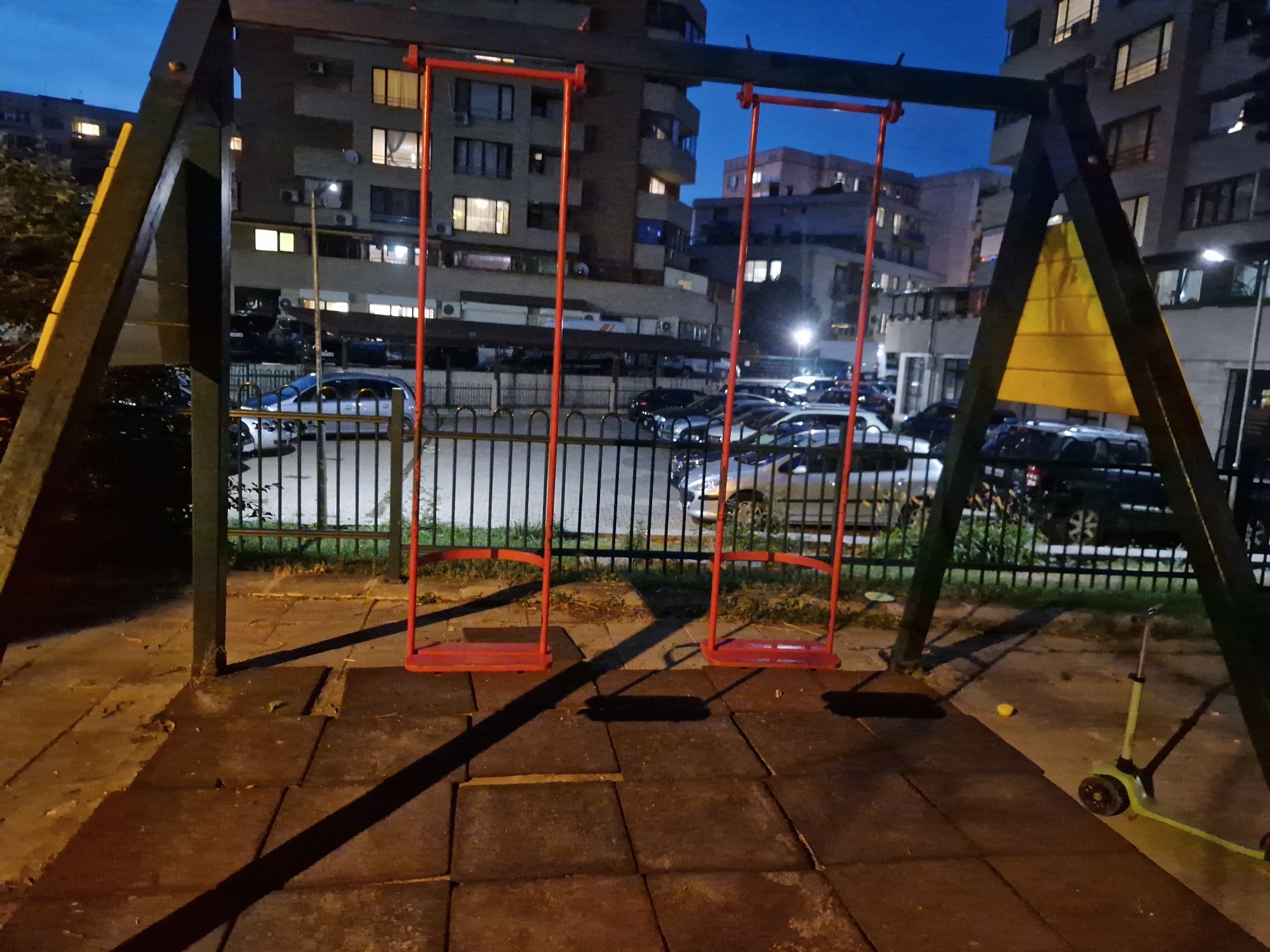 The image depicts a playground swing set with two red swings, located in Sofia, Bulgaria. The swing set is made of dark-colored wood and metal. It is situated on a surface of dark, square rubber tiles. In the background, there are apartment buildings with lit windows, suggesting it is evening or night. A black metal fence separates the playground from a parking area with several parked cars. A yellow scooter is visible on the right side of the image, near the swing set. The lighting in the scene is a combination of artificial light from the buildings and streetlights, casting long shadows on the playground.