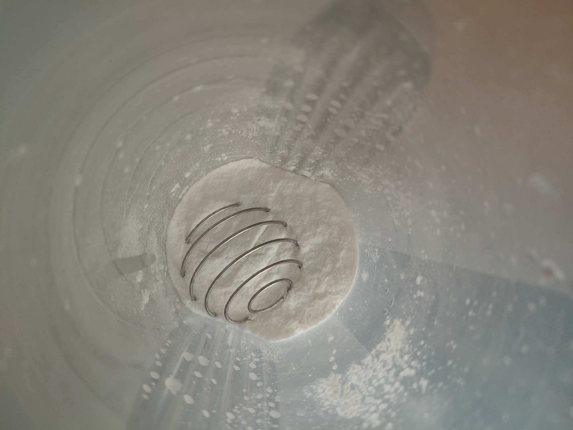 The image presents a top-down view into a light-colored, cylindrical container, resembling a shaker bottle. At the bottom of this container, a distinct mound of white powdery substance is visible. Resting within this powder is a spherical metal whisk, characterized by its coiled, spring-like design.
The inner walls of the container are smooth and curved, with various specks and residues of the white powder clinging to them, particularly noticeable on the lower sections. This residue suggests that the container has either been previously used for mixing or that the powder has recently been poured in. The lighting appears soft and even, illuminating the contents without harsh shadows.
The scene depicts the preparation phase for mixing a powdered drink or ingredient, with the whisk in place, ready to combine the powder with a liquid. There are no people visible, and no discernible text or branding within the frame. The focus is sharp on the whisk and the powder, while the upper parts of the container walls show a slight blur, emphasizing the contents at the bottom.