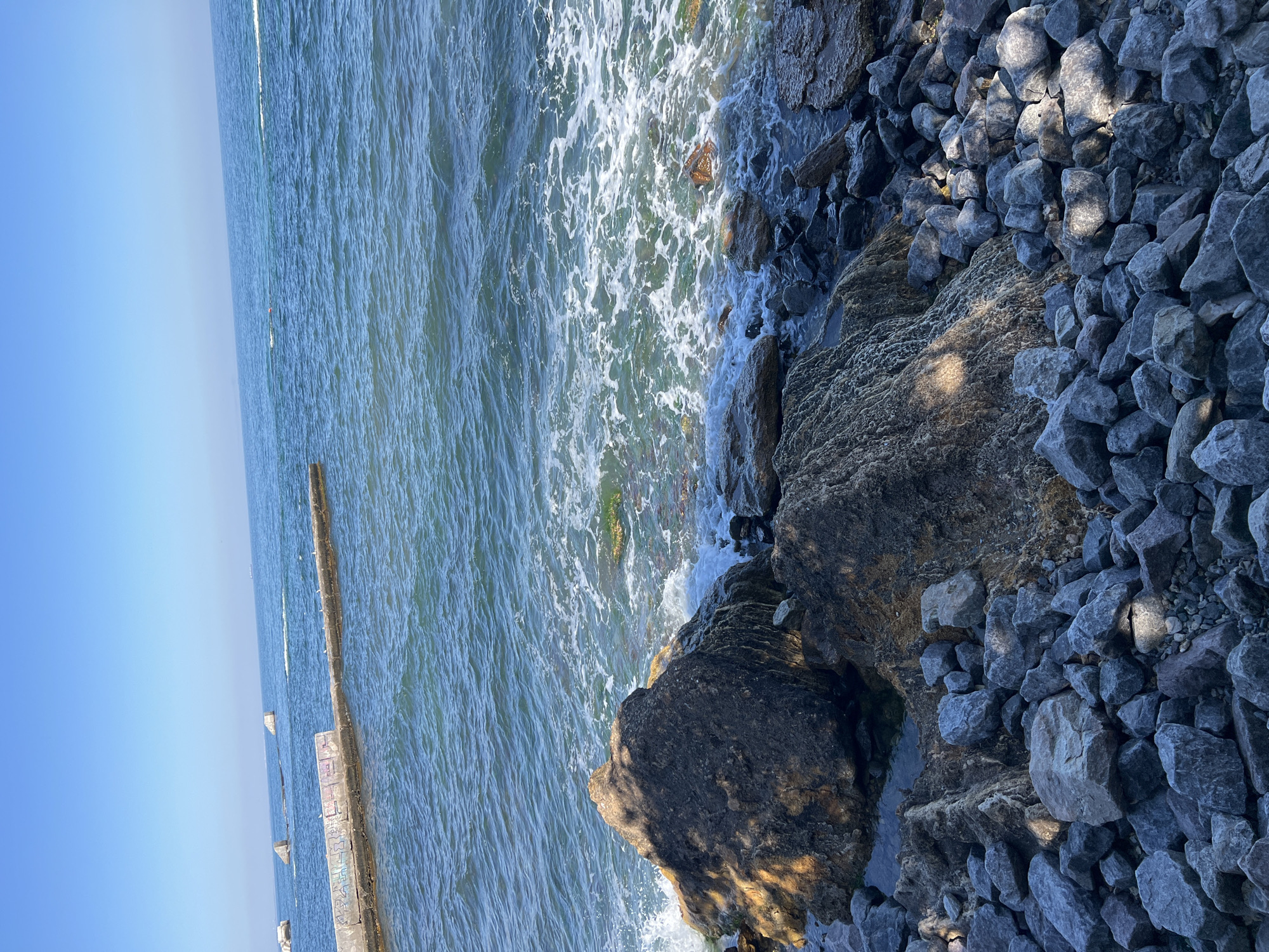 A rocky coastline with the sea and a pier is depicted. The sky is clear and blue. In the foreground, there are many large and small rocks. Some of the rocks in the foreground are large and dark, with areas of light hitting them. The water is a deep blue-green with white foam from the waves crashing against the rocks. In the middle ground, there is a concrete pier with graffiti on it and several birds sitting on it. In the background, the sea stretches to the horizon, with a few distant objects that may be buoys or submerged structures. The overall scene is one of a natural coastal environment with some man-made elements. The location is the Odesa coast.
