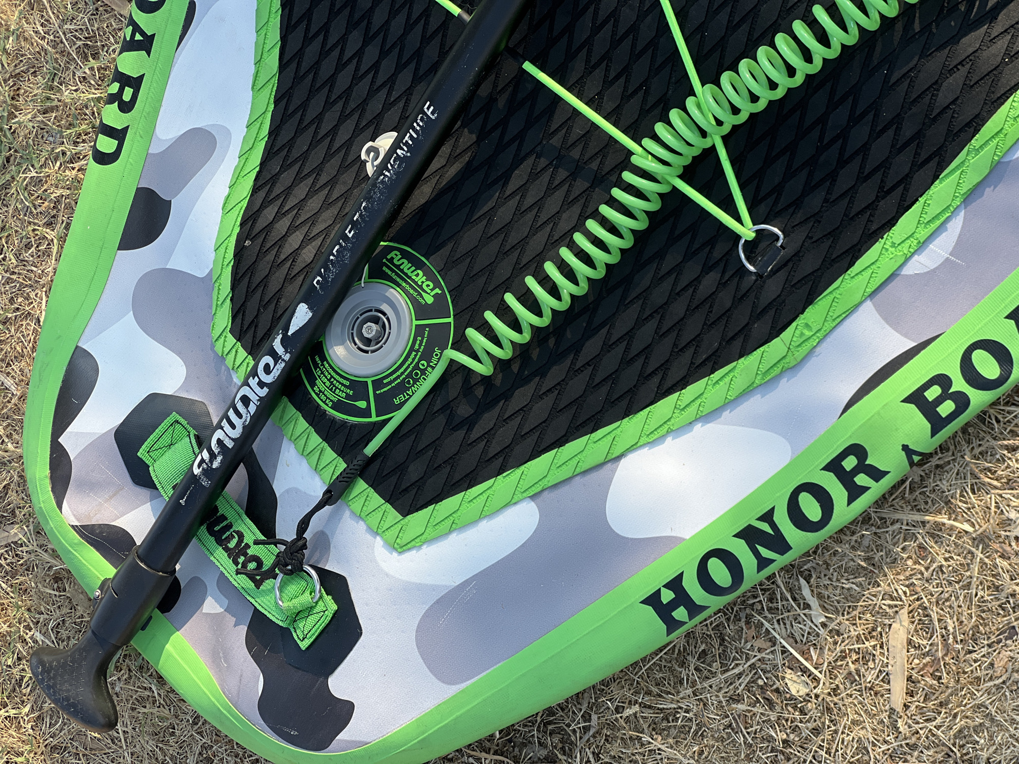 An overhead close-up shot shows a black paddle with a green handle and a black coil leash attached to a green and gray camouflage inflatable paddleboard. The paddleboard has black grip tape and green trim. The words "HONOR BOARD" are visible in black lettering on the green trim. The scene is set on dry, sun-baked grass, suggesting an outdoor location in Antalya, Turkey, possibly a park or beach area during a dry season. There are no people visible in the image.