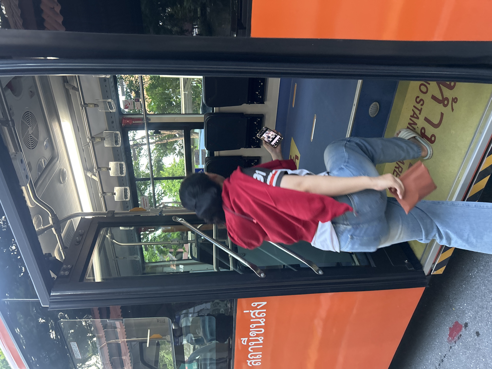 A young Thai woman is captured from behind, stepping onto a bus in Tha Khlong Town Municipality, Thailand. She is wearing light blue denim jeans, a white t-shirt with black and red stripes on the sleeves, and a red hooded sweatshirt draped over her shoulders. In her right hand, she holds a smartphone, and in her left, a small brown pouch or wallet. The bus is orange with Thai writing on its side. The bus interior features blue flooring with yellow and red warning stripes near the entrance, and rows of black seats. Overhead, metal handrails are visible. The scene appears to be outdoors, with trees and buildings visible through the bus windows.