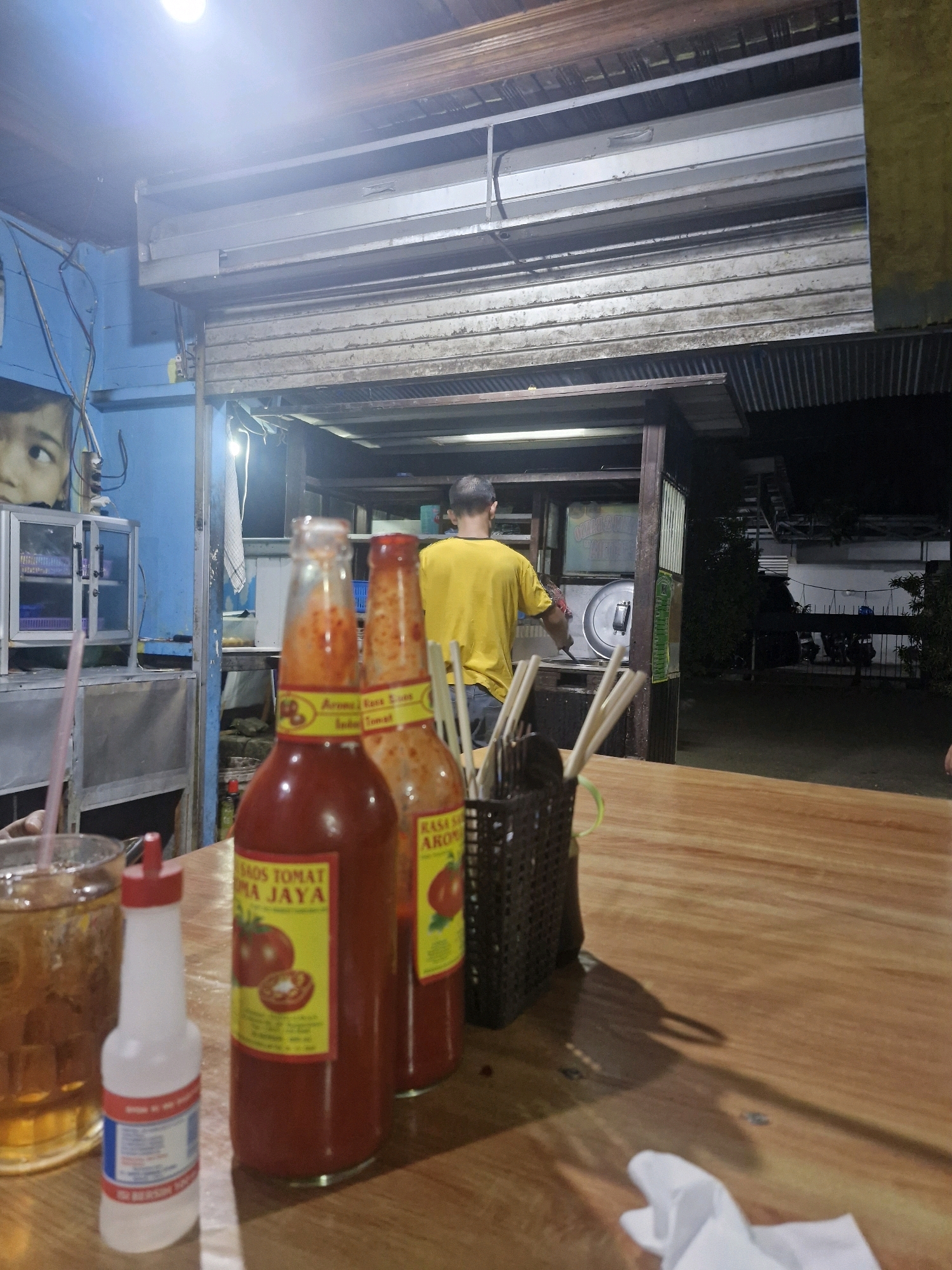 In Rantau, Indonesia, an outdoor food stall is captured at night. The foreground shows a wooden table with two large glass bottles of red tomato sauce, labeled "SOS TOMAT", and a small plastic bottle with a red cap, likely a condiment. A glass of iced tea with a straw sits to the left. In the midground, a person wearing a yellow t-shirt is seen from behind, cooking at a counter. Behind the counter, a metal shutter is partially rolled up, revealing a dimly lit interior. The background shows a dark outdoor area with trees and what appear to be parked motorcycles. The overall scene depicts a typical, casual dining environment at a local eatery.