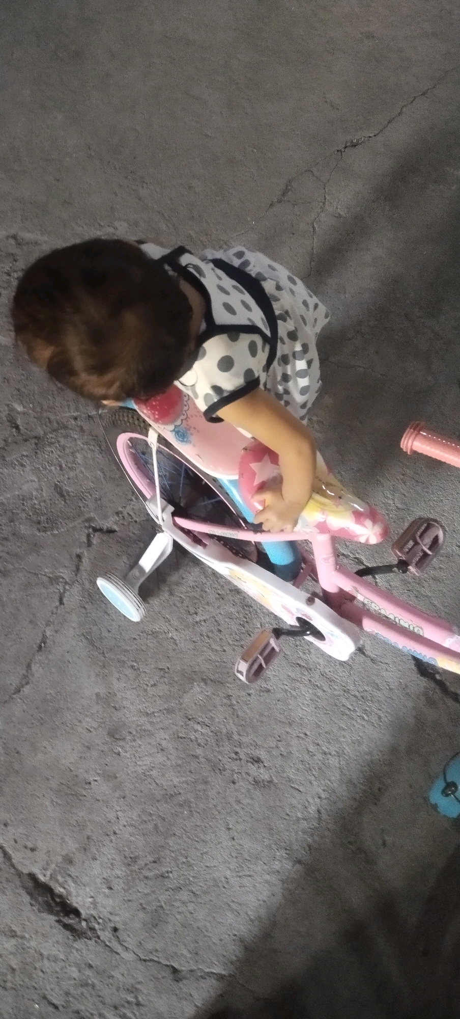 A young child wearing a polka-dot dress is leaning over a pink children's bicycle on a cracked concrete surface in Daleman, Indonesia. The child's head is down, obscuring their face, and their hands are on the bicycle's seat. The bicycle has training wheels and decorative decals. To the right of the bicycle, a pink handlebar is visible. The overall environment appears to be an outdoor area with a rough, gray concrete ground.
