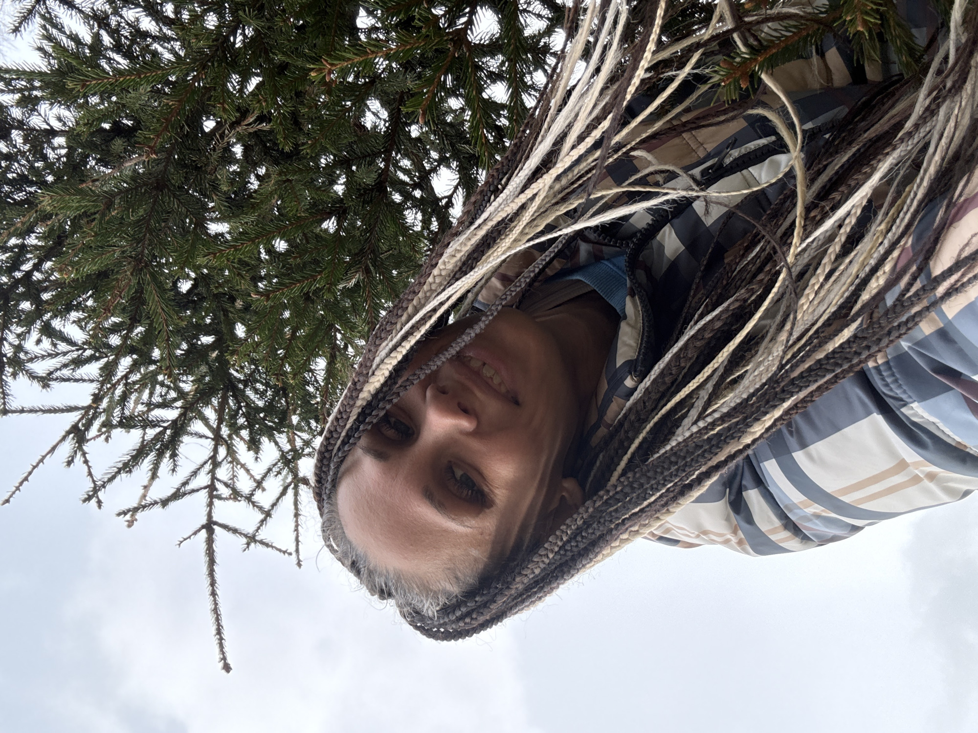 The image presents a low-angle portrait of a person, likely an adult female, captured with a rotated orientation. She is smiling gently and looking directly at the camera, her head positioned in the lower right of the frame, angled as if gazing upwards. Her notable feature is long, multi-toned braided hair, featuring shades of dark brown, black, and light blonde, cascading towards the left. She is wearing a jacket adorned with a plaid or striped pattern in muted tones of blue, beige, and white.
Above and to the left of the person, the dense, dark green branches and needles of a coniferous tree, possibly a spruce or fir, dominate the upper and central portions of the background. Through and beyond these branches, and filling the remaining background, is an overcast sky, uniformly bright and appearing white or light gray. This indicates a daytime setting with cloudy weather. The scene suggests an outdoor natural environment, such as a forest or park, located in Rusanovo, Russia. No text is visible in the image. The person's expression conveys a calm and pleasant mood.