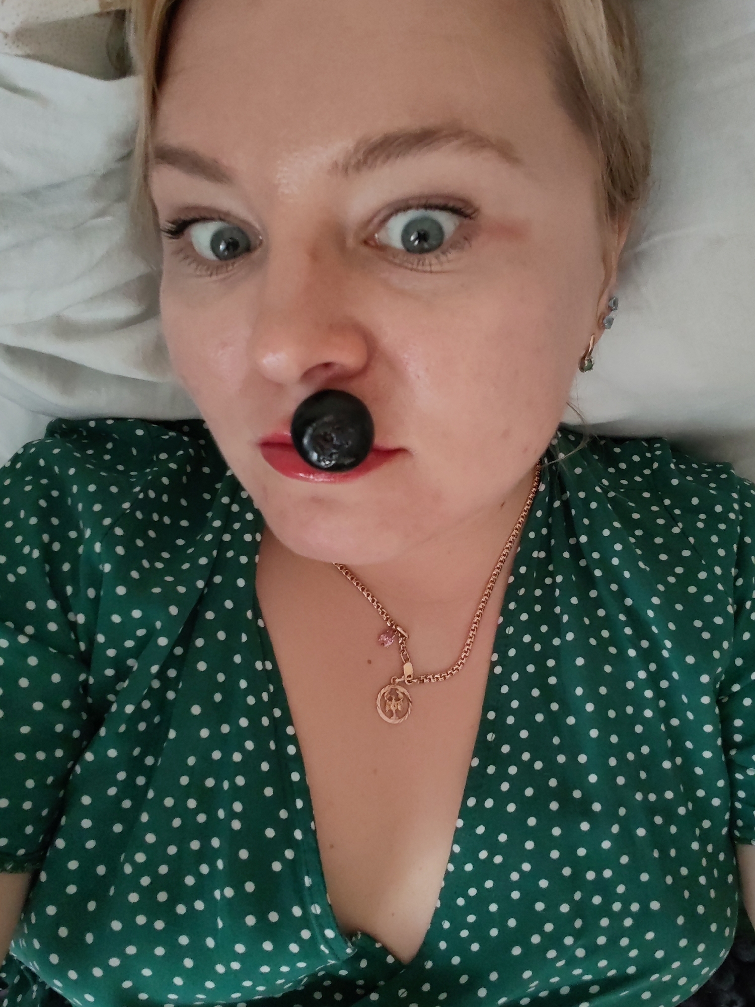 A close-up, high-angle portrait captures a light-skinned individual, likely female, with blonde hair, lying on a white surface and looking directly up at the camera. Her eyes are wide and appear light blue-grey, accentuated by dark eyelashes. Her lips are colored with glossy red or pink lipstick, and a large, dark, spherical object, possibly a grape, is playfully held between her upper lip and nose.
She is wearing a green dress patterned with small white polka dots, featuring a V-neckline. Around her neck is a gold-colored chain necklace with a round pendant displaying an indiscernible emblem and a smaller pink charm. In her visible left ear, she wears multiple gold-colored earrings, one appearing to have a light blue or green stone. The expression conveyed is one of surprise or playfulness. The setting is indoors, with soft white fabric suggesting a bed or cushion, located in Izhevsk, Russia. There is no visible text in the image.