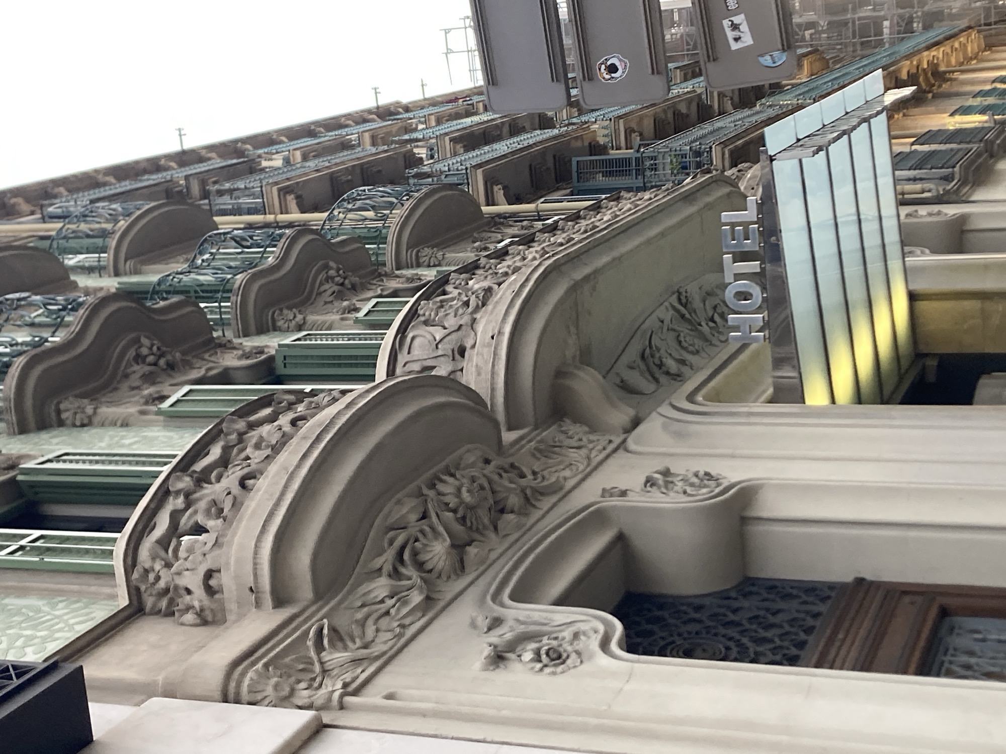The image shows an exterior view of a hotel in Barcelona, Spain. The architecture features intricate decorative stonework on the building's facade, including balconies with ornate railings and sculpted floral motifs. A prominent "HOTEL" sign is visible above the entrance, which is partially covered by a modern, glass awning. The building has multiple stories with green-shuttered windows. In the background, scaffolding suggests ongoing construction or renovation work. The sky is overcast.