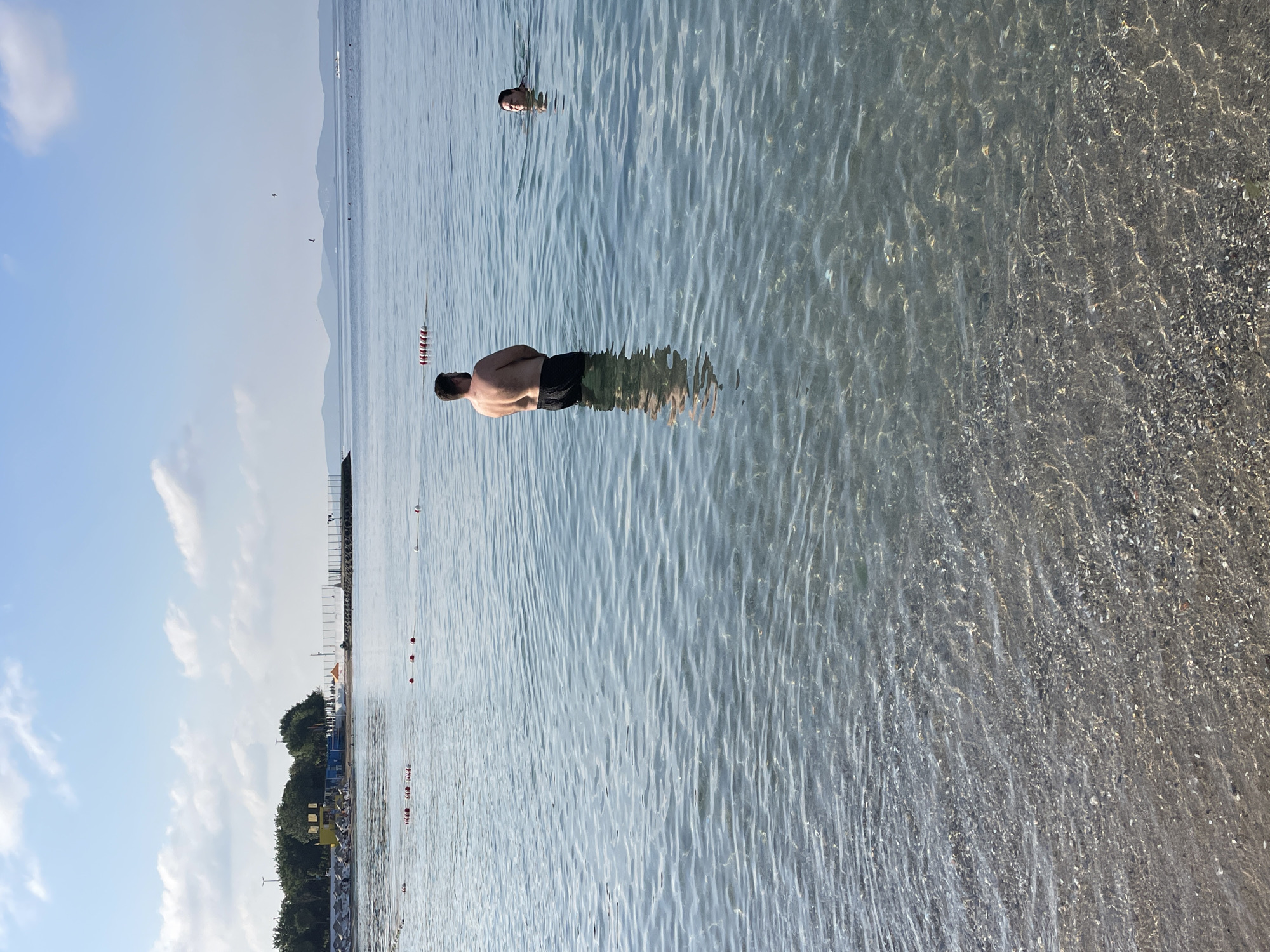 This image depicts a serene view of the sea at Moda Beach in Kadıköy, Istanbul, Turkey. Two individuals are present in the water. A man, seen from the back, stands waist-deep, wearing black swim trunks. Further out, a woman is swimming, with only her head and shoulders visible above the water. The water is clear, allowing the seabed to be seen. In the background, a shoreline is visible with trees, beach umbrellas, and a pier. The sky is bright blue with scattered clouds, and a chain of buoys stretches across the water. The overall scene conveys a sense of a calm, sunny day at the beach.