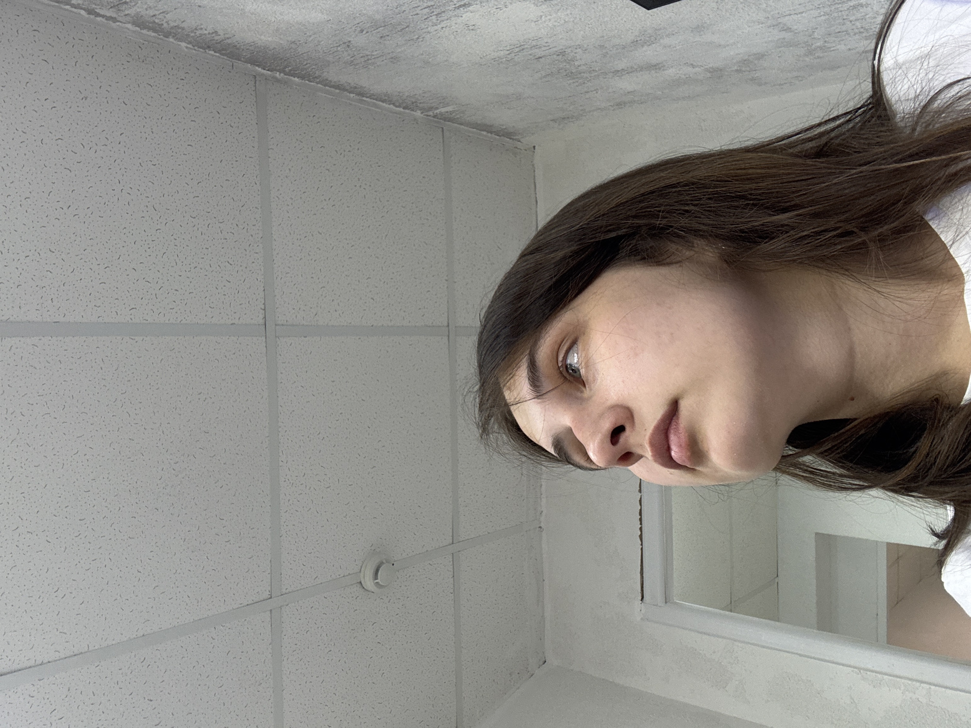 A woman with brown hair and fair skin is visible, partially reflected in a mirror. The surrounding environment includes a room with a suspended ceiling consisting of square tiles. The specific location within the city of Chaikovsky, Russia, cannot be determined from the visual information provided. The scene consists of a self-portrait. The woman appears to be looking at the camera.