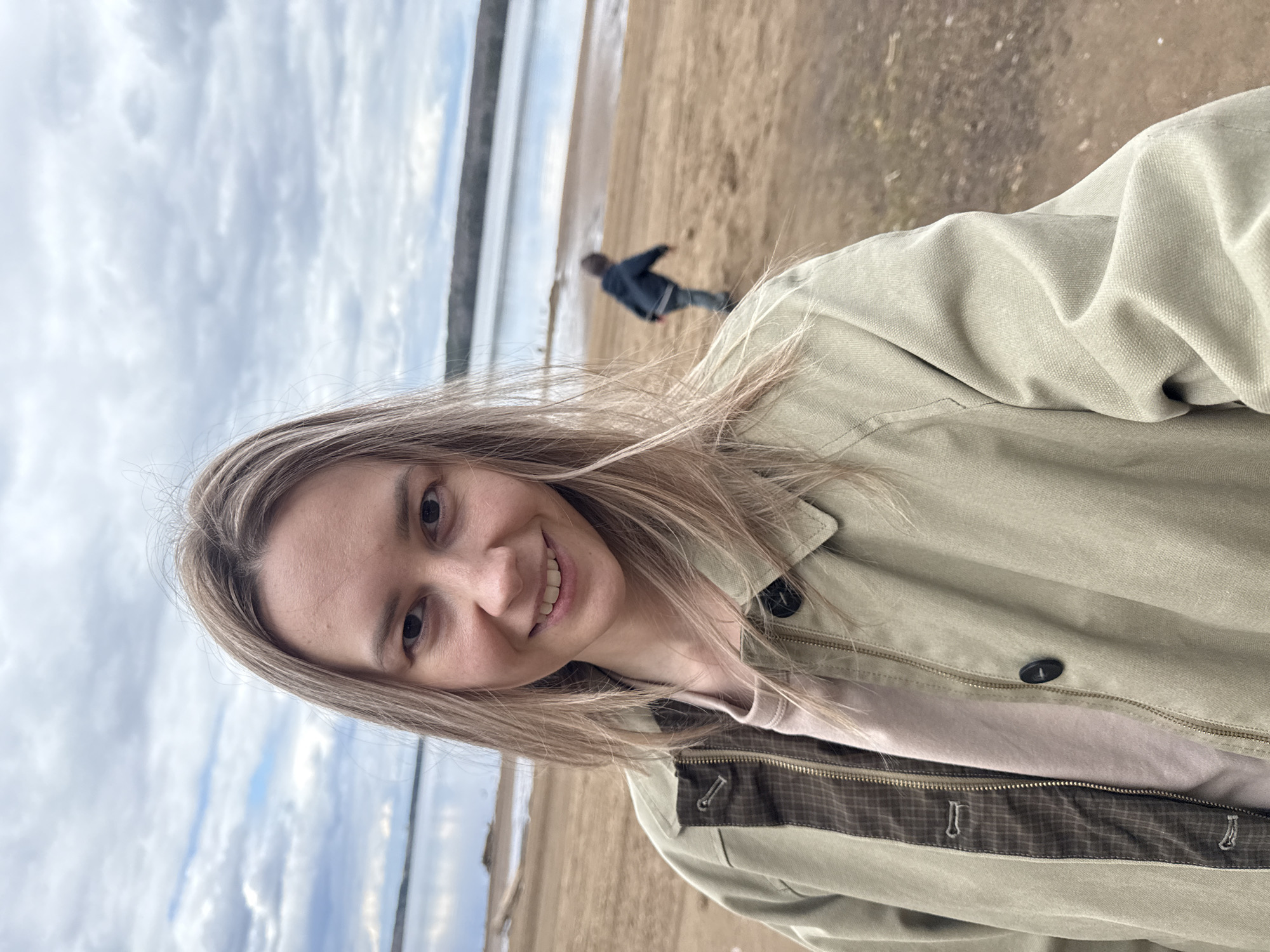 The image is a slightly low-angle selfie of a smiling woman, taken outdoors on a cloudy day. When viewed upright, the woman's face and upper torso are prominent on the right side of the frame. She has long, light brown or blonde hair and is looking directly at the camera with a happy expression. She wears a textured light olive-green jacket, open at the front to reveal a light pink or beige top. Visible details on her jacket include a black button, a zipper, and a dark brown plaid-patterned trim along the inner lining or lapel.
Behind the woman, to her left, lies a wide, sandy or muddy shoreline next to a calm body of water, likely a lake or wide river, consistent with the Chaikovsky, Russia location context. The sky is overcast with soft, diffused clouds. In the mid-ground, a blurry figure of a person, possibly a child, dressed in dark clothing, is seen walking or running on the sandy bank, moving away from the camera. The overall scene suggests a leisurely outing on a cool, cloudy day. No visible text is present.