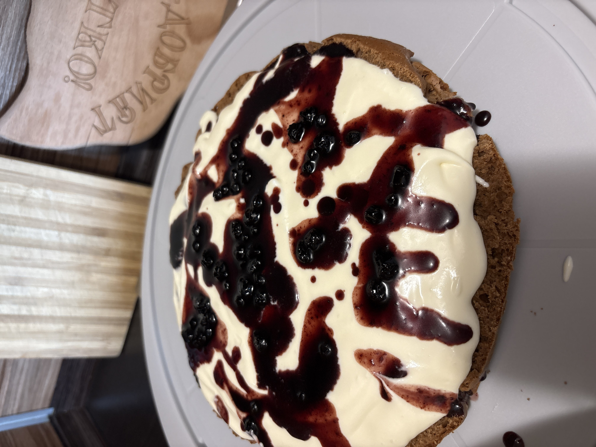 A close-up, eye-level shot shows a round cake topped with white frosting, blueberries, and a dark berry syrup. The cake is on a gray cake stand. In the background, there is a wooden cutting board with text carved into it, and a portion of a dark countertop. The lighting is natural and appears to be from overhead.