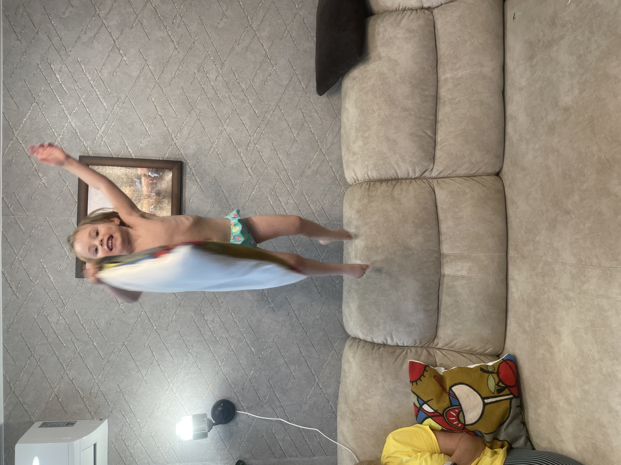 In this photo, a young blonde girl is captured mid-air, jumping with a pillow. She's wearing a colorful swimsuit, and her arms are outstretched, suggesting an energetic leap. The girl's face shows excitement and joy.
The background features a patterned grey wallpaper and a section of a large, light-colored sectional sofa. A framed picture hangs on the wall behind the girl. To the right, a portion of the sofa is visible, with a decorative pillow featuring a bold, abstract design. Another person, wearing a yellow shirt and striped pants, is partially visible lying down on the sofa, their face obscured by the decorative pillow.
A white appliance, possibly a humidifier or air purifier, is visible in the lower left corner, next to a black lamp that emits a bright light. The overall scene suggests a playful indoor environment, likely a living room. The time of day is not explicitly indicated, but the artificial lighting suggests it could be evening or a dimly lit interior space. The presence of a sofa and the domestic setting point towards a home environment in Vladivostok, Russia.