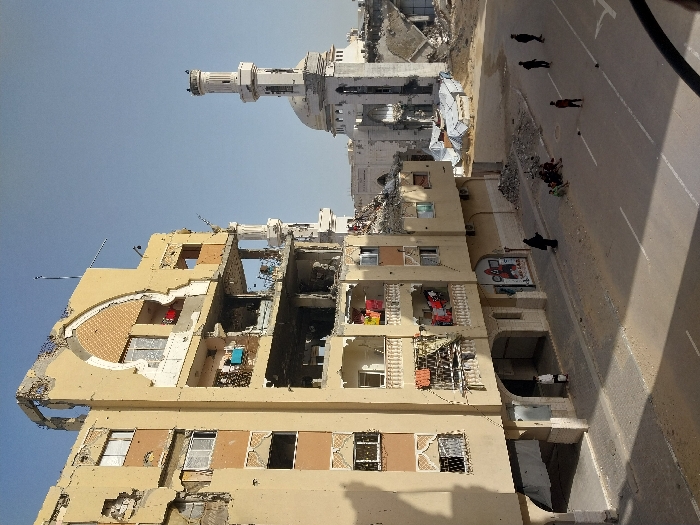 The image depicts a scene of extensive urban destruction in Khan Yunis, Palestinian Territory, under a clear daylight sky. In the foreground, a multi-story, tan-colored concrete building exhibits severe structural damage, with large sections of its facade and upper floors missing, exposing interior rooms and twisted rebar. Balconies with ornate railings remain partially intact on the lower and middle floors, some displaying hanging laundry. The ground level features arched doorways, one occupied by an individual in a white garment, and a storefront adorned with a red-masked figure mural.
In the mid-ground, a damaged large white mosque with a distinctive minaret and partially collapsed dome is visible. Debris piles are scattered on the ground between the mosque and the street. The street itself is paved with asphalt, marked by white lines, and has adjacent sidewalks.
Several individuals are present: one person in a dark abaya stands on the sidewalk near the damaged building, while a group of approximately five individuals, appearing to be children or youths, are seated on the adjacent sidewalk. Three additional individuals, also appearing as youths, stand scattered on the asphalt street further to the right. The overall environment conveys a significant impact event with ongoing human presence amidst the destruction.