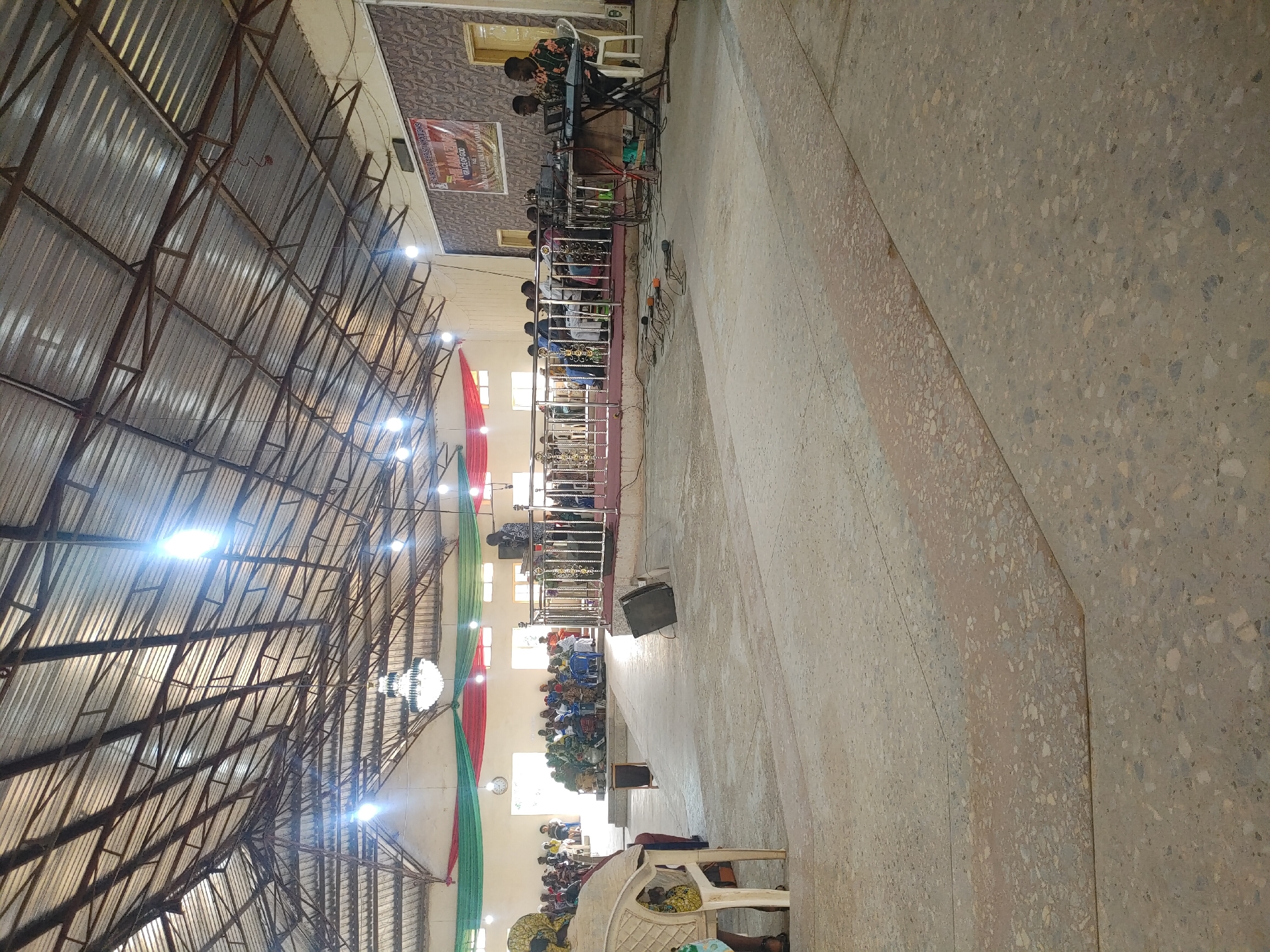 The image shows an indoor gathering in what appears to be a church or community hall in Jos, Nigeria. The space has a high, corrugated metal roof supported by a metal framework. Colorful drapes in red and green are suspended from the ceiling. A chandelier hangs in the center of the hall, and several bare light bulbs are also visible.
In the background, a group of people are seated, facing a raised platform. On the platform, a woman is standing and speaking, likely a preacher or speaker. To the right of the platform, three men are seated behind a table with musical equipment, including keyboards and microphones. One man is playing a keyboard.
In the foreground, a woman is seated in a chair, facing away from the camera. The floor is made of concrete and has a speckled appearance. There are also several microphones and cables on the floor in front of the stage. A sign with text is visible on the wall behind the musicians, but the text is too blurry to read. The overall impression is of a religious service or community event.