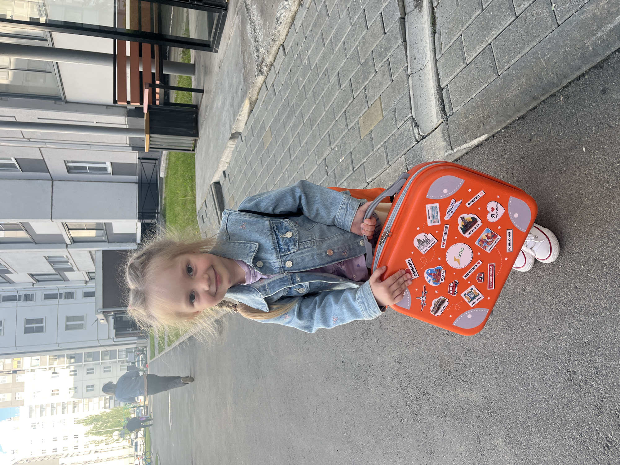 A young girl with blonde hair pulled into two braids, wearing a denim jacket and white sneakers, stands on an asphalt sidewalk in front of a residential building. She is holding an orange suitcase adorned with travel-themed stickers. The suitcase features images of airplanes, landmarks, and text in Russian. In the background, a woman is walking away from the camera, and other people are visible in the distance. The environment appears to be a typical urban setting with paved walkways, grassy areas, and apartment buildings. The presence of stickers on the suitcase and the general atmosphere suggest a playful or travel-related context. The text on the suitcase includes "Сибирь", "Москва", "Екатеринбург", "Челябинск", and "Я Путешествую" (I travel).