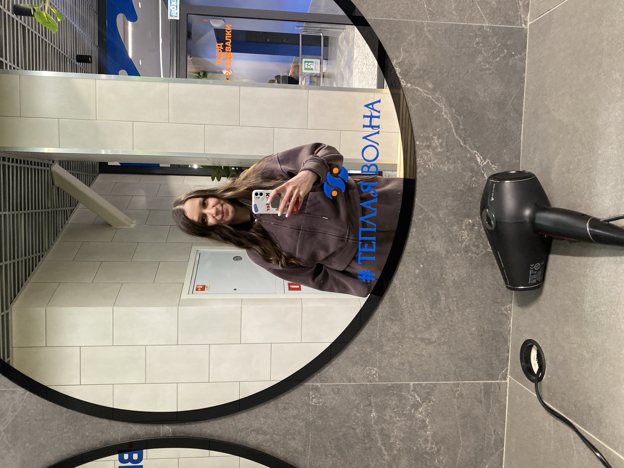 A young woman is taking a selfie in a round mirror. The mirror's edge is black. Reflected in the mirror are various elements including interior walls, door signage in Cyrillic, and a ventilation grate. The woman is wearing a brown hooded sweatshirt and holding a phone with a decorated case. The mirror features blue Cyrillic text and logos. To the right of the mirror, on a gray tiled wall, is a black hair dryer and its power cord disappearing into a wall outlet. This is located in the water park "Теплая Волна" in Votkinsk, Russia.