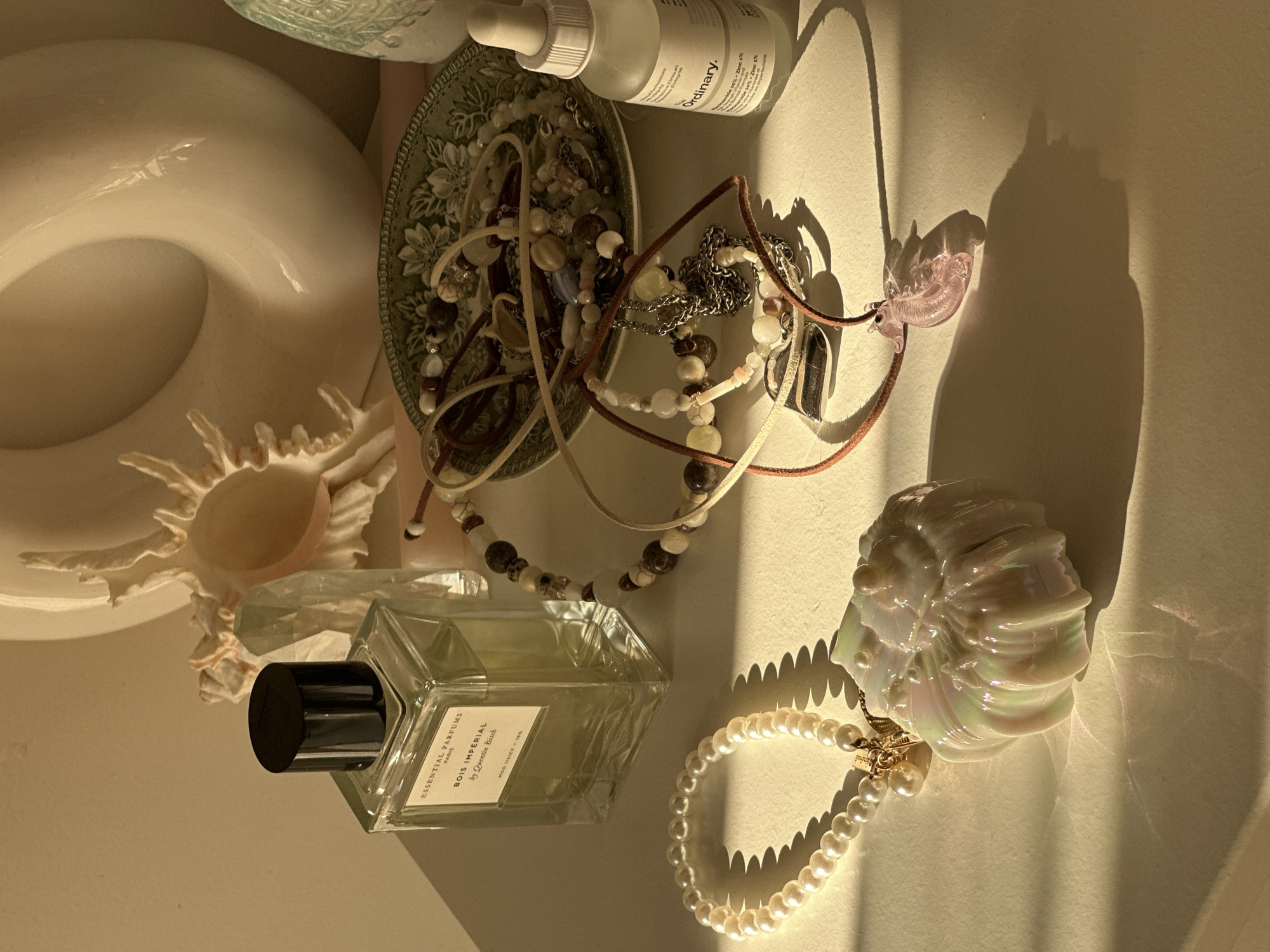 The image is a high-angle shot featuring a collection of objects arranged on a surface, likely a table or shelf. The lighting is warm, possibly from natural sunlight, casting long shadows.
Objects include:
- A white ceramic doughnut-shaped vase or object.
- A seashell.
- A small green floral-patterned plate filled with tangled jewelry.
- A bottle of "The Ordinary" brand skincare product.
- Several strands of beaded necklaces, including one with a pink glass pendant.
- A rectangular glass bottle of "Essential Parfums Bois Imperial" perfume.
- A string of pearls.
- A shell-shaped object with a pearlescent finish.
The scene does not contain any indication of people or location in Bar, Montenegro. The setup appears to be a carefully arranged still life.
