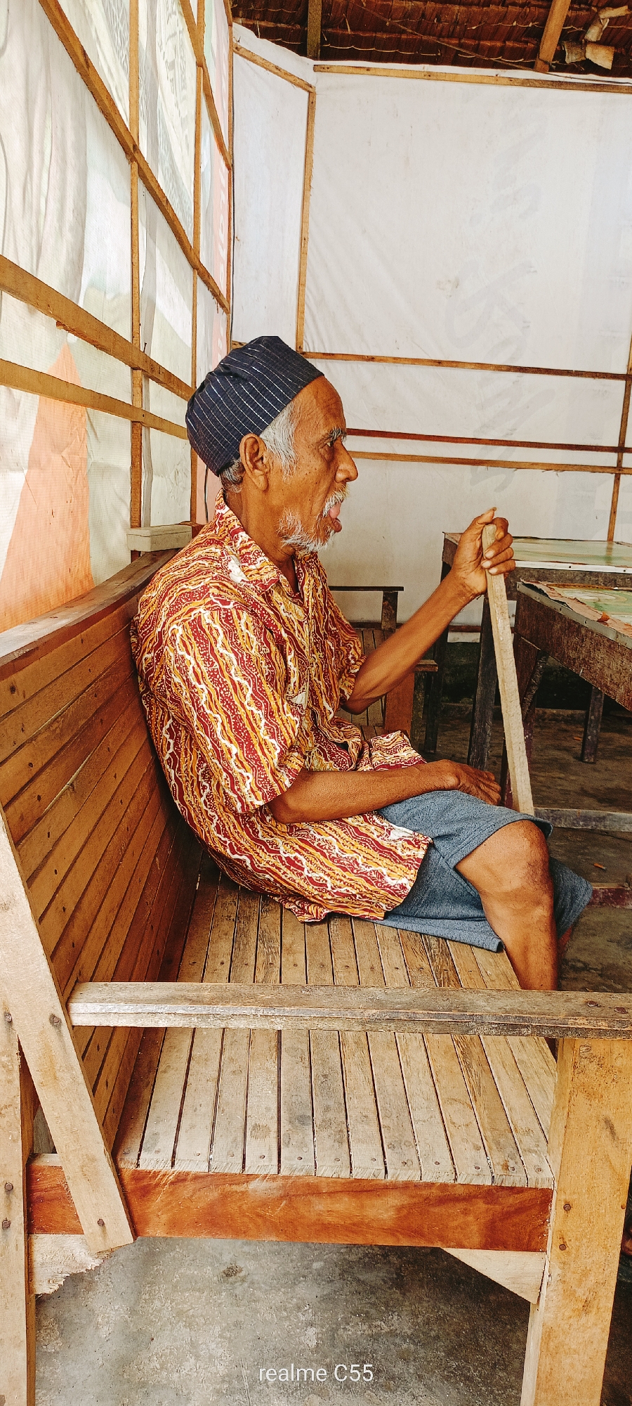 An elderly man sits on a wooden bench, his body angled to the right, with his head turned and mouth slightly open in conversation or speech. He wears a dark blue cap with thin white stripes and a colorful, patterned short-sleeved shirt in red, yellow, and white. His legs are clad in grey shorts, and his right hand rests on a wooden stick. The bench is constructed from light-colored wooden planks, with visible nails and slight wear. Behind the man, a table with a patterned surface is partially visible. The background consists of draped fabric, likely white or light-colored, supported by wooden beams, and a thatched roof. The overall environment appears to be an open-air structure. The image was captured in Lhokseumawe, Indonesia, as indicated by the "realme C55" watermark, which is a mobile phone model.