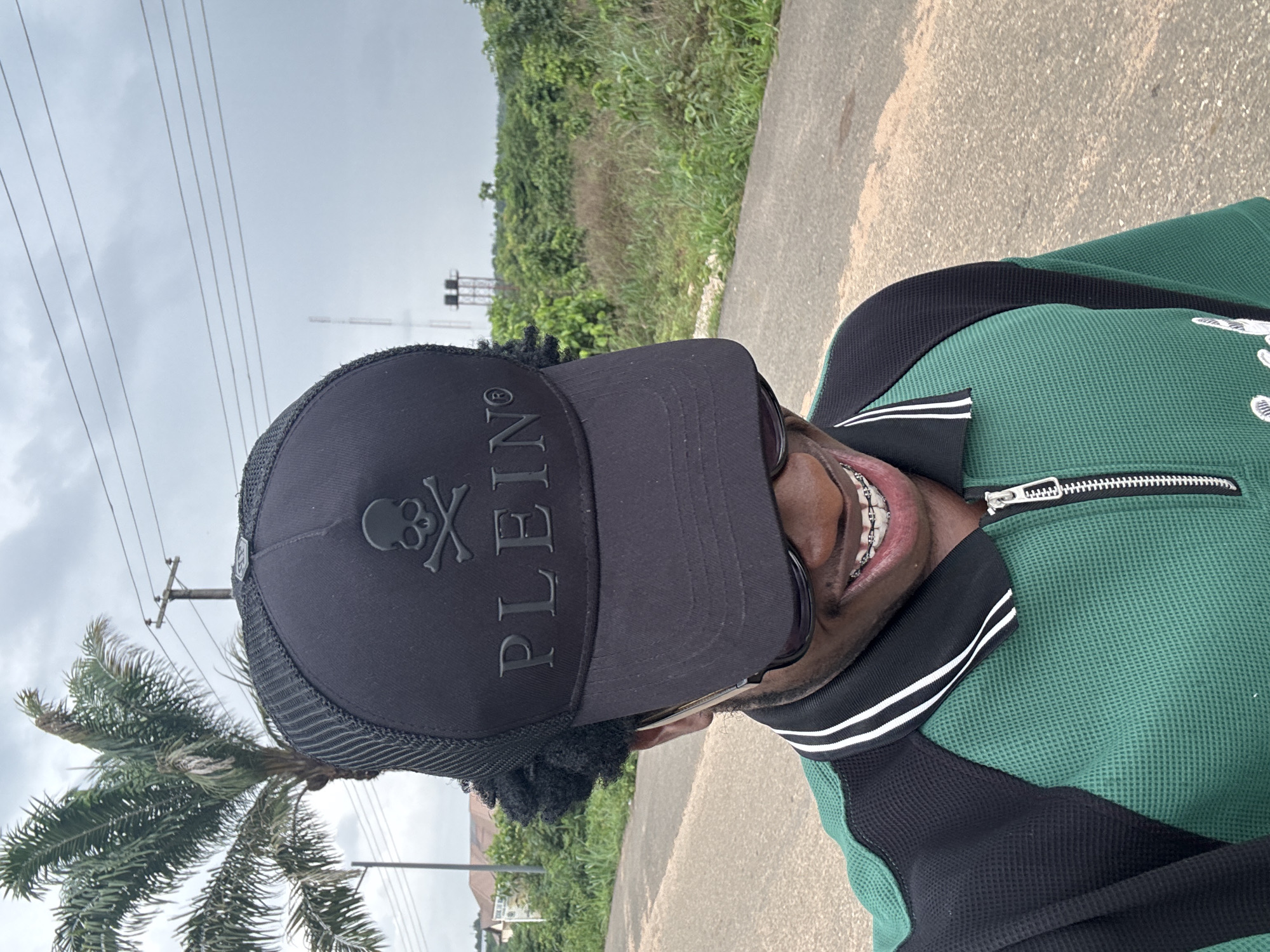 A person, presumably male, is captured in a close-up, selfie-style photograph in Ihiagwa, Nigeria. The individual is wearing a black cap emblazoned with a skull and crossbones above the word "PLEIN". Sunglasses are visible, and braces are present on the individual's teeth. The person is wearing a green and black zip-up jacket. The background features a street with lush greenery and overcast skies with power lines. A palm tree is visible on the left side of the image.