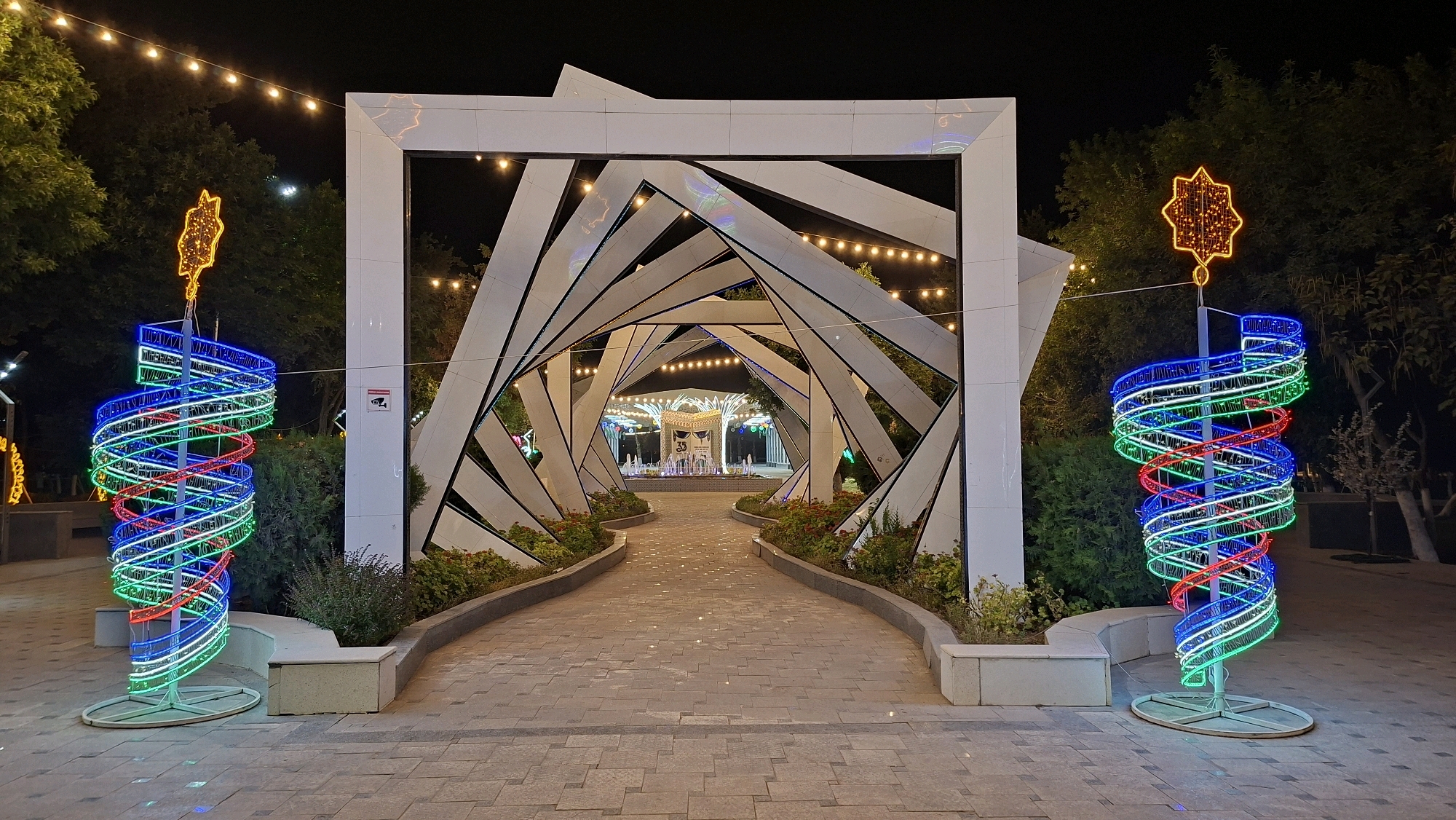 The image depicts an outdoor scene at night, likely a public square or park in Navoiy City, Uzbekistan. The foreground features a paved pathway made of large, rectangular stone tiles with some scattered reflective lights. On either side of the pathway, flanking the entrance to an archway, are tall, spiral-shaped light installations. These installations consist of a central pole wrapped in coiled LED lights of blue, green, and red, topped with an illuminated starburst design.
The central focus is a modern, abstract archway structure composed of intersecting white, angular planes. This archway frames a pathway leading deeper into the scene, where more lights are visible. Beyond the archway, a carousel or similar festive attraction with colorful lights and a fountain in front of it can be seen in the distance. String lights are hung overhead across the scene, illuminating the path and surrounding trees. Lush green bushes and trees form the backdrop on either side of the pathway and behind the archway. A security camera is visible mounted on a white pillar to the right of the archway. The overall ambiance is festive and decorative, suggesting a nighttime event or holiday decoration.