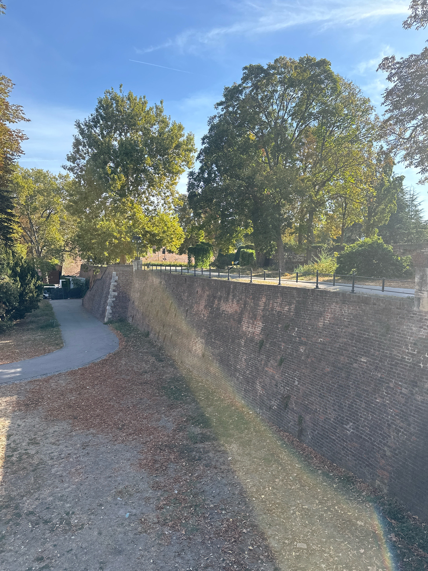 The image depicts a section of the Belgrade Fortress walls in Belgrade, Serbia, on a clear, sunny day. A paved pathway runs alongside the ancient brick wall, which is overgrown with some moss and vegetation. The ground to the left of the pathway is covered in fallen leaves, indicating an autumn season. Tall trees with green foliage are visible behind the wall, casting shadows. The sky is a bright blue with a faint contrail from an aircraft. A subtle rainbow-like light refraction is visible on the lower right portion of the image, likely caused by the sun's rays interacting with the camera lens or atmospheric conditions. No people are present in the scene.