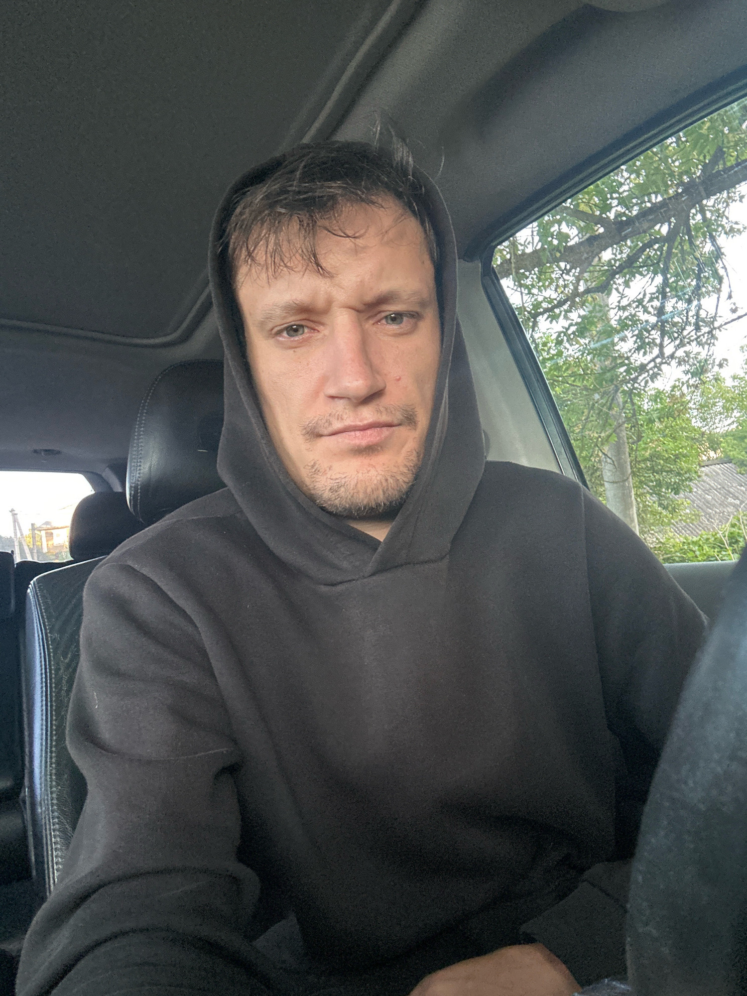 A man wearing a black hoodie is seated in the driver's seat of a car. The car's sunroof is open, and trees and buildings are visible through the open window. The man is looking directly at the camera, and his facial expression is neutral. The interior of the car is dark, but the exterior appears to be a daytime scene in a rural or suburban area. The image is taken from a selfie perspective.