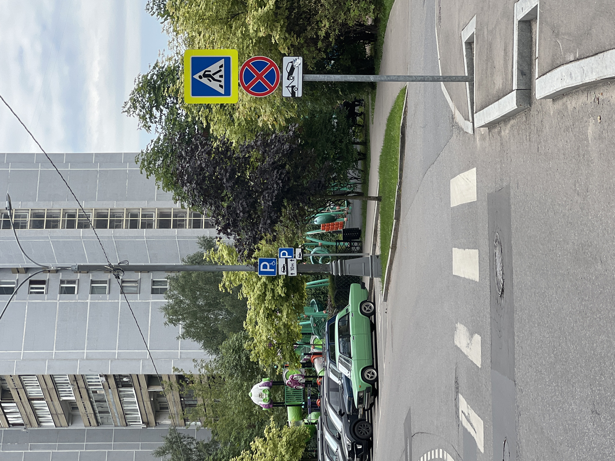 The image captures a residential street scene in Moscow, Russia, on an overcast day. In the background, a tall, grey, multi-story apartment building with numerous balconies and windows dominates the skyline. In the mid-ground, a children's playground with colorful structures is visible, surrounded by lush green trees. Several cars are parked along the street, including a light green sedan prominently featured in the foreground. Traffic signs are mounted on a metal pole: a blue square sign with a white pedestrian silhouette indicates a crosswalk, a red circle with a blue background and a red cross signifies no stopping, and a rectangular sign below depicts a tow truck towing a car. A pedestrian crosswalk is clearly marked on the asphalt road. The environment appears to be a typical urban residential area, with a focus on community amenities like a playground and accessible parking.
