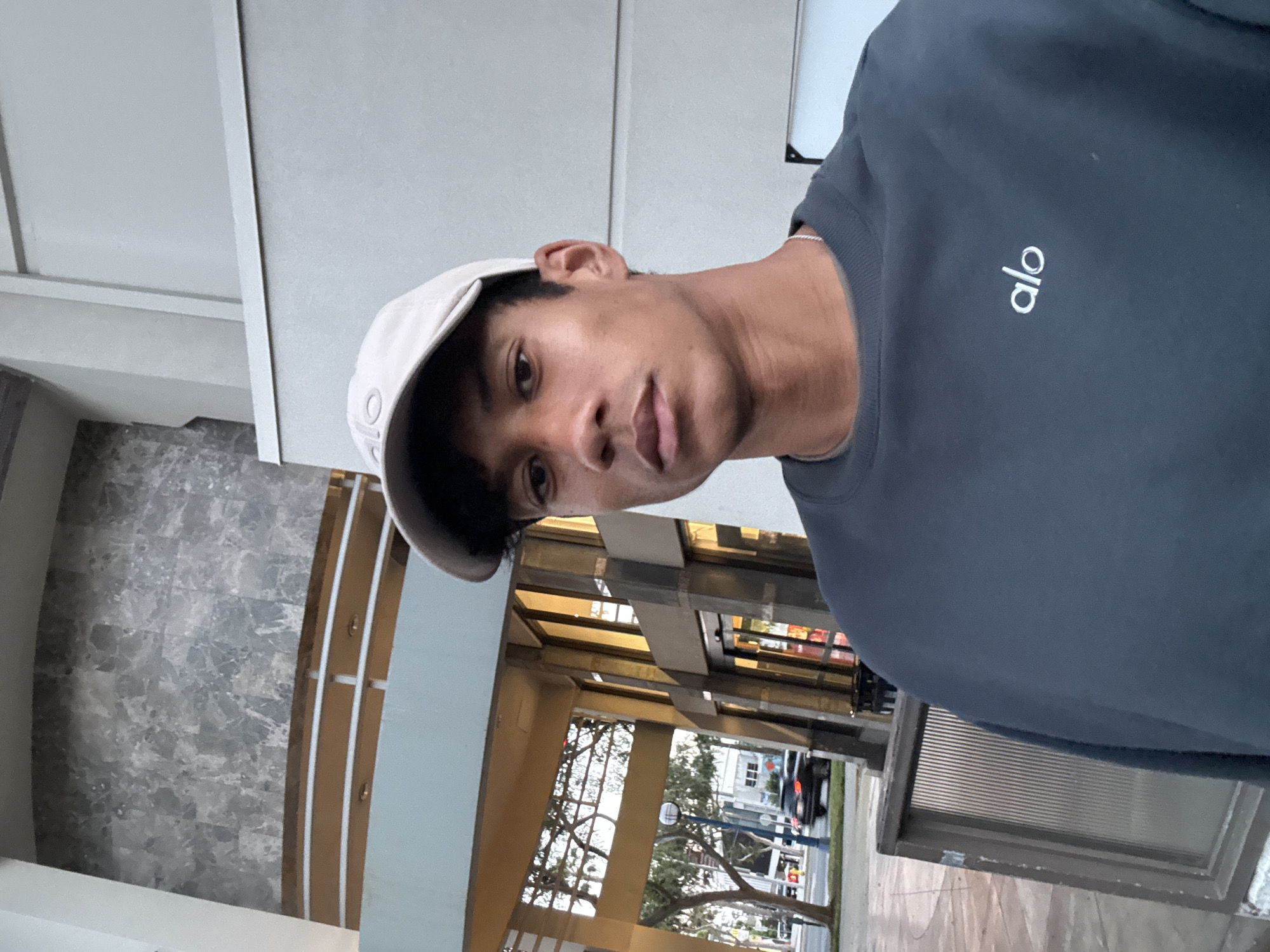 A medium shot presents a person wearing a dark gray "Alo" sweatshirt and a light-colored baseball cap, positioned in front of an exterior architectural structure with curved elements and a gray stone accent wall. The background includes trees, a building with large windows, and streetlights. A glimpse of a vehicle and what appears to be storefronts further defines the urban environment. The location is West Hollywood, United States of America.