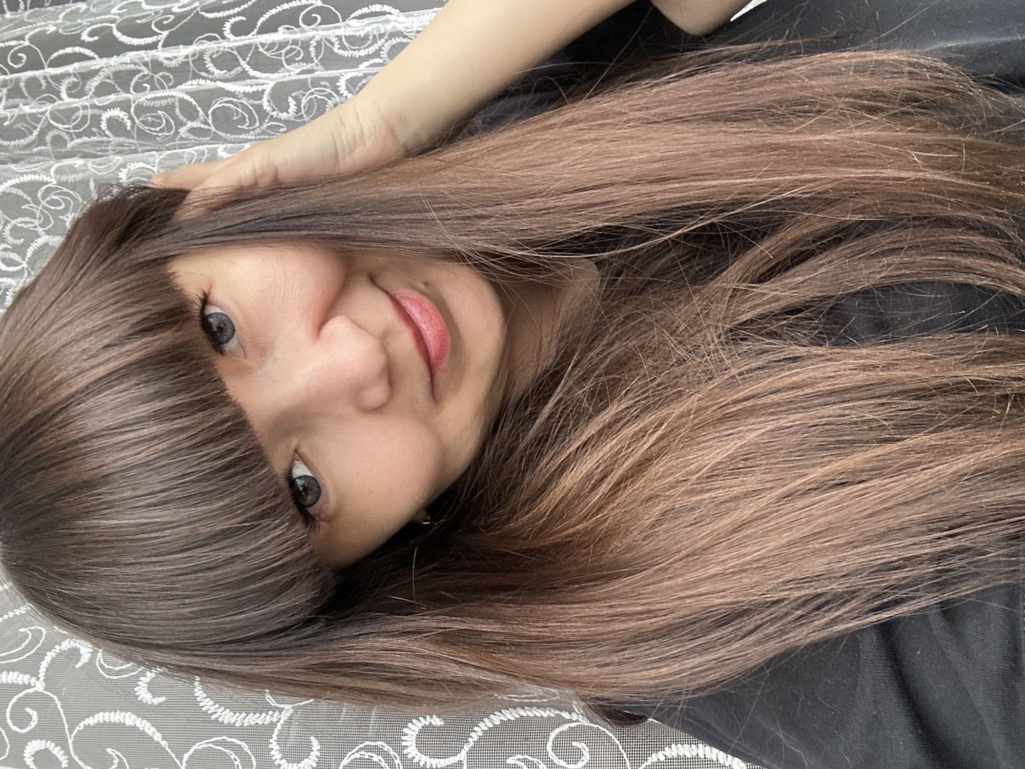 A person with long, straight brown hair and bangs is looking at the camera with a slight smile. The person's hair is partially obscuring their face, and they have light skin and gray eyes. The person is wearing a black shirt, and the background consists of a patterned white lace or mesh material. The location is likely indoors but cannot be pinpointed to Chaikovsky, Russia based on the provided image.