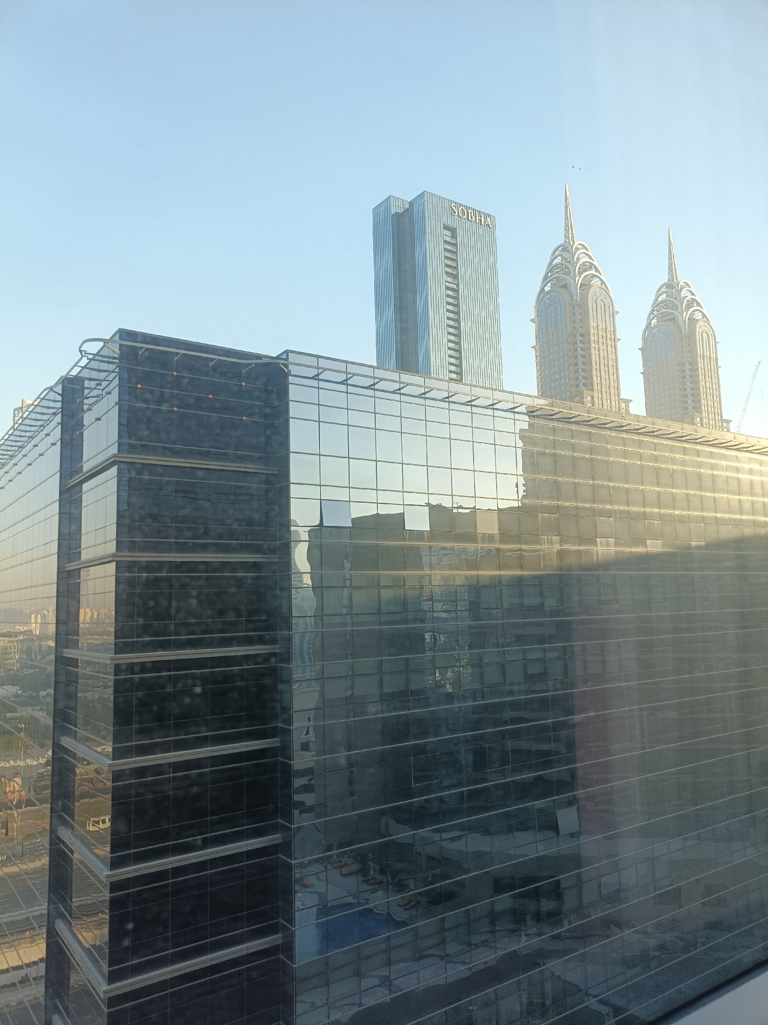 This image captures a view of the Sobha Realty building and the Opus building in Dubai, United Arab Emirates. The Sobha Realty building is a tall, modern skyscraper with a glass facade. The Opus building is a distinctive architectural design, featuring two towers with unique spires. Both buildings are set against a clear blue sky. In the foreground, there is a glass-enclosed building that reflects the surrounding environment. The scene appears to be from a high vantage point, looking out over the city. The overall impression is one of urban development and contemporary architecture.