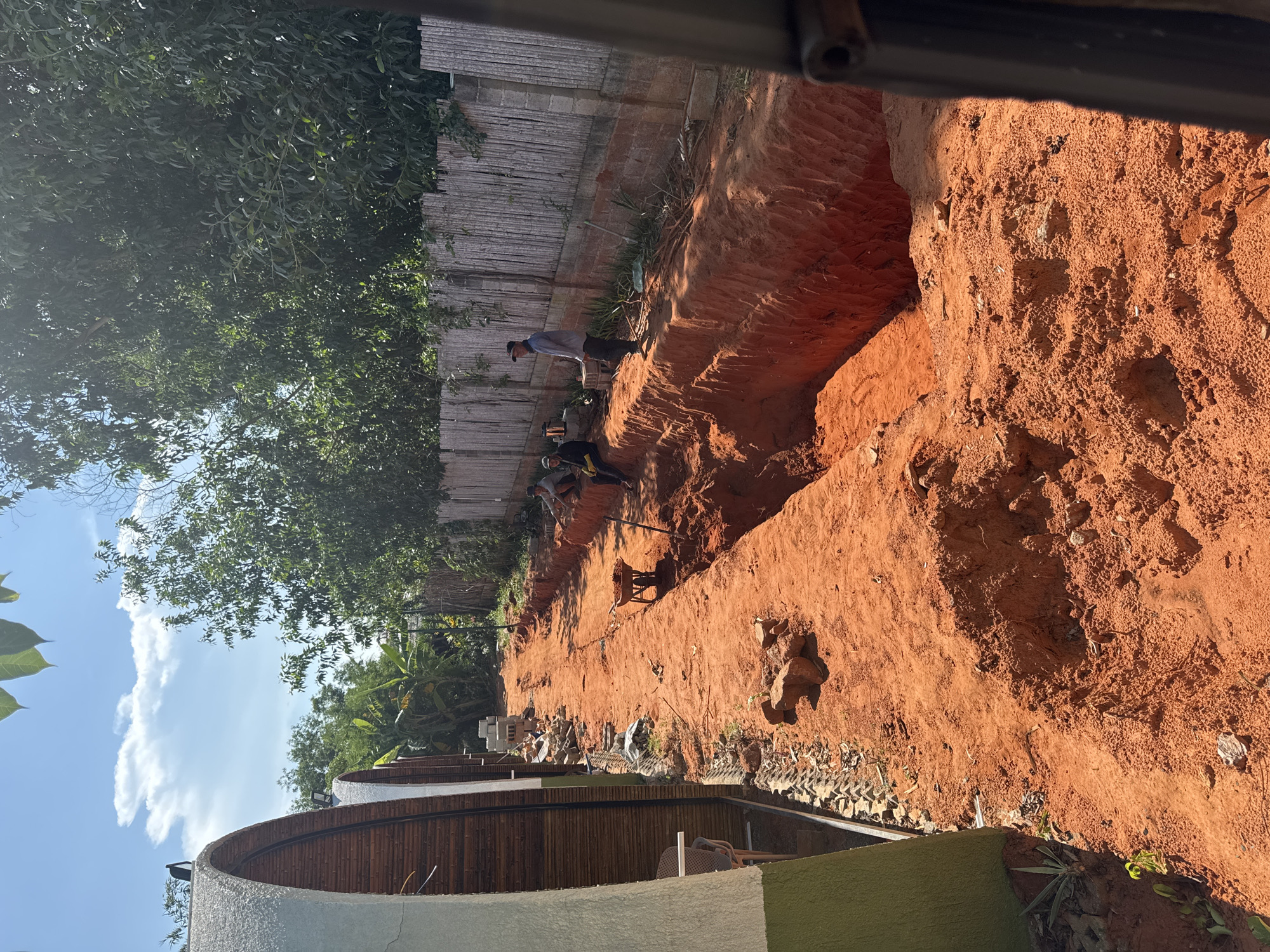 The image depicts a construction site in Mũi Né, Vietnam, with a deep trench dug into the reddish-orange soil. The trench appears to be an ongoing excavation, with visible layers of earth. Several individuals are present, engaged in construction activities. One person, wearing a light-colored shirt and a baseball cap, stands at the edge of the trench, observing the work. Another individual, seated, is visible further down the trench. A wheelbarrow loaded with soil is positioned near the trench, suggesting the transport of excavated material. To the left of the trench, a row of distinctive dome-shaped structures, likely accommodations, are visible. These structures feature a white exterior and bamboo-like siding. A wooden fence, composed of vertical slats, runs along the right side of the trench, bordering the property. Vegetation, including trees and some banana plants, is abundant in the background, indicating a tropical or subtropical environment. The sky is blue with scattered white clouds, and the sunlight suggests it is a clear day. The overall scene conveys a sense of ongoing development and construction work in a scenic, natural setting.