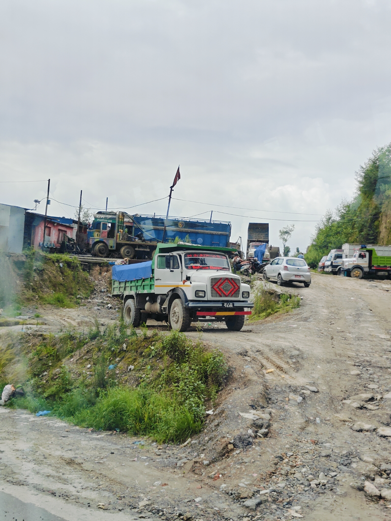 The image depicts a scene on a rural, unpaved road in Chandragiri, Nepal, with several trucks and cars present. A white dump truck with a green bed and a blue tarp covering its cargo is in the foreground, facing slightly left. Behind it, a blue and white truck with decorative elements is parked. Further back, more trucks and a silver car are visible on the road, which is uneven and appears to be under construction or in disrepair. The environment is hilly, with green vegetation growing on the slopes. The sky is overcast with a light gray hue. Several power lines and poles are visible in the background, suggesting a settlement nearby. A red flag is planted on a pole in the distance. The overall impression is of a working area with heavy vehicle traffic on a challenging terrain.
