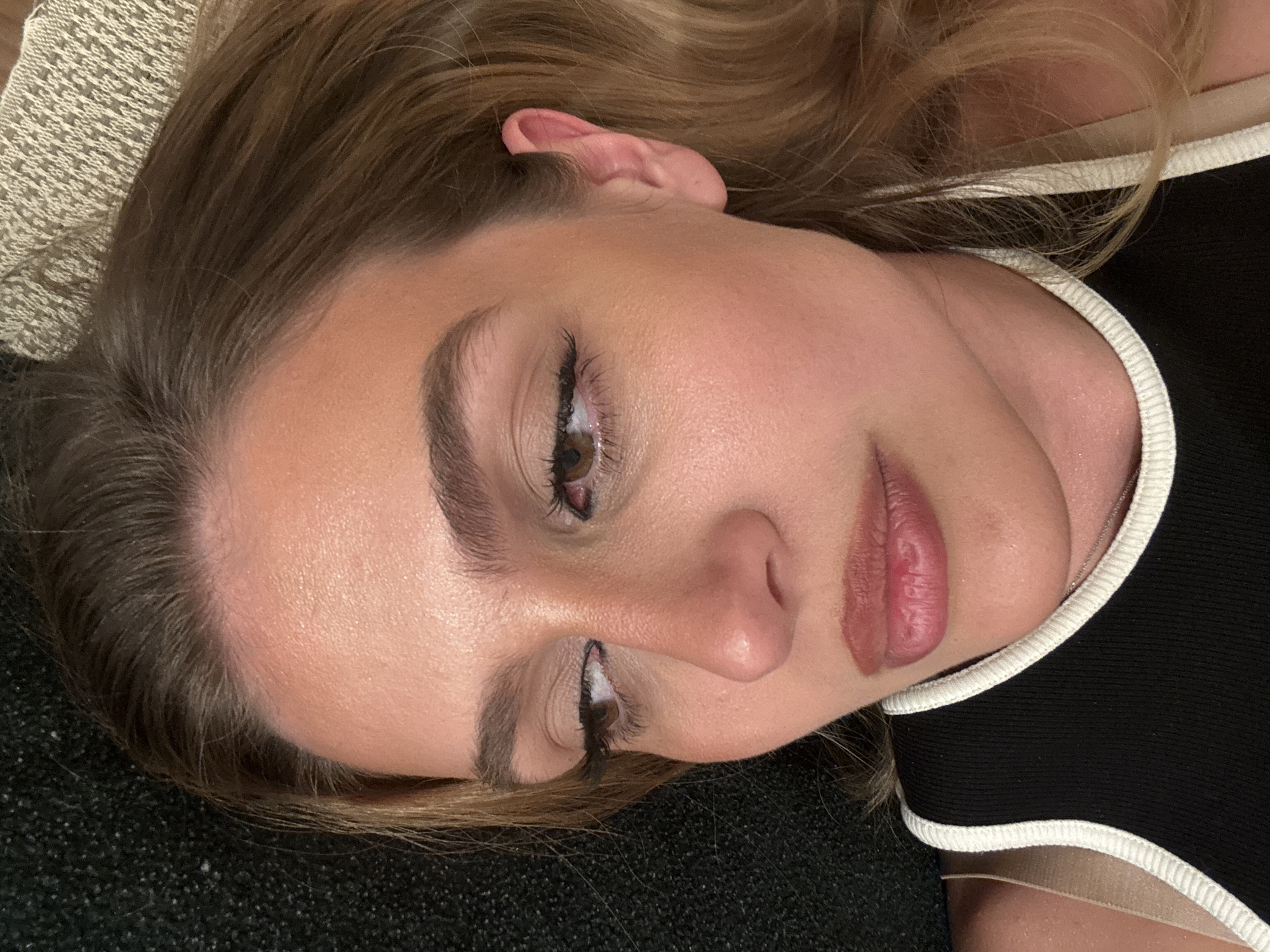 A close-up, high-angle shot captures a person lying down, looking upward with a relaxed and neutral expression. The subject's head is tilted, showing the right side of their face and a portion of their left ear. Their eyes are open, revealing brown irises framed by dark eyeliner and mascara. The eyebrows are well-defined. The nose is prominent, and the lips are full, appearing to have a natural-toned lip color.
The person's hair, a mix of light brown and blonde hues, is loosely spread around their head, suggesting a relaxed pose. They are wearing a black top with a white trim along the neckline and shoulder strap. A delicate chain necklace is barely visible near the collarbone. The background consists of a dark, textured fabric, possibly a blanket or cushion, on which the person's head rests. Another lighter, patterned fabric is visible to the upper left of their head, possibly another cushion or part of the bedding. The lighting is soft and even, highlighting the subject's features without harsh shadows. No text is visible in the image.