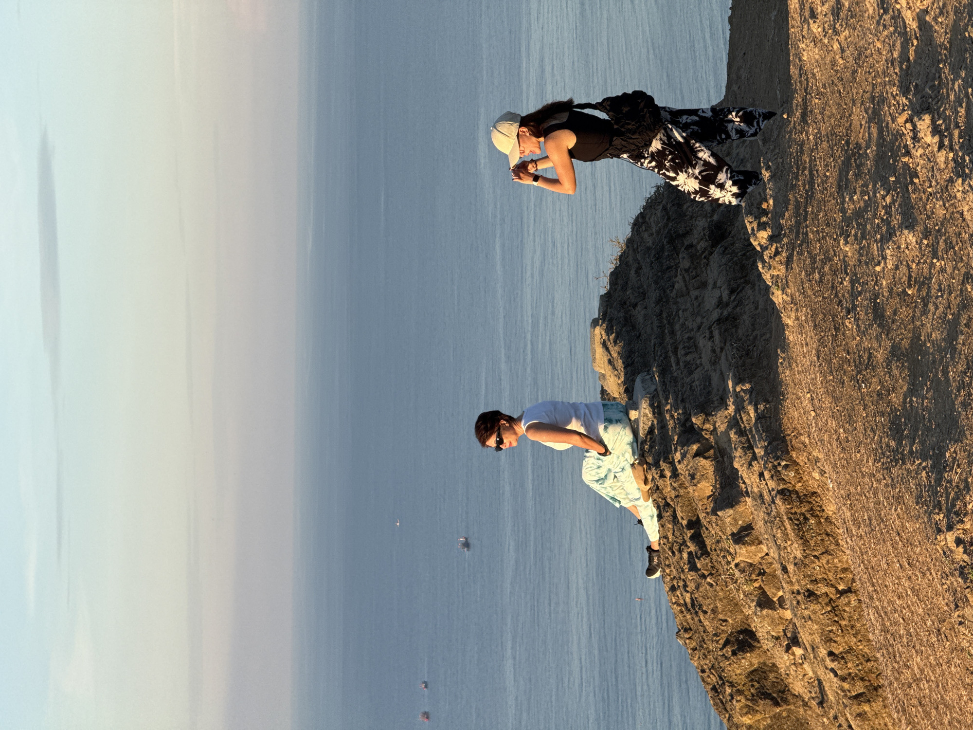 The image captures a scenic view from a cliff overlooking the ocean. Two women are present on the rocky terrain. One woman, seated on a rock, wears sunglasses and a white tank top with patterned pants. She gazes towards the ocean. The second woman stands nearby, holding a camera and appearing to take a photograph. She is wearing a black tank top, a baseball cap, and patterned pants. In the distance, several boats are visible on the calm blue water, and the sky displays a gradient of soft pastel colors. The location is the Lý Sơn Special Administrative Region of Vietnam.
