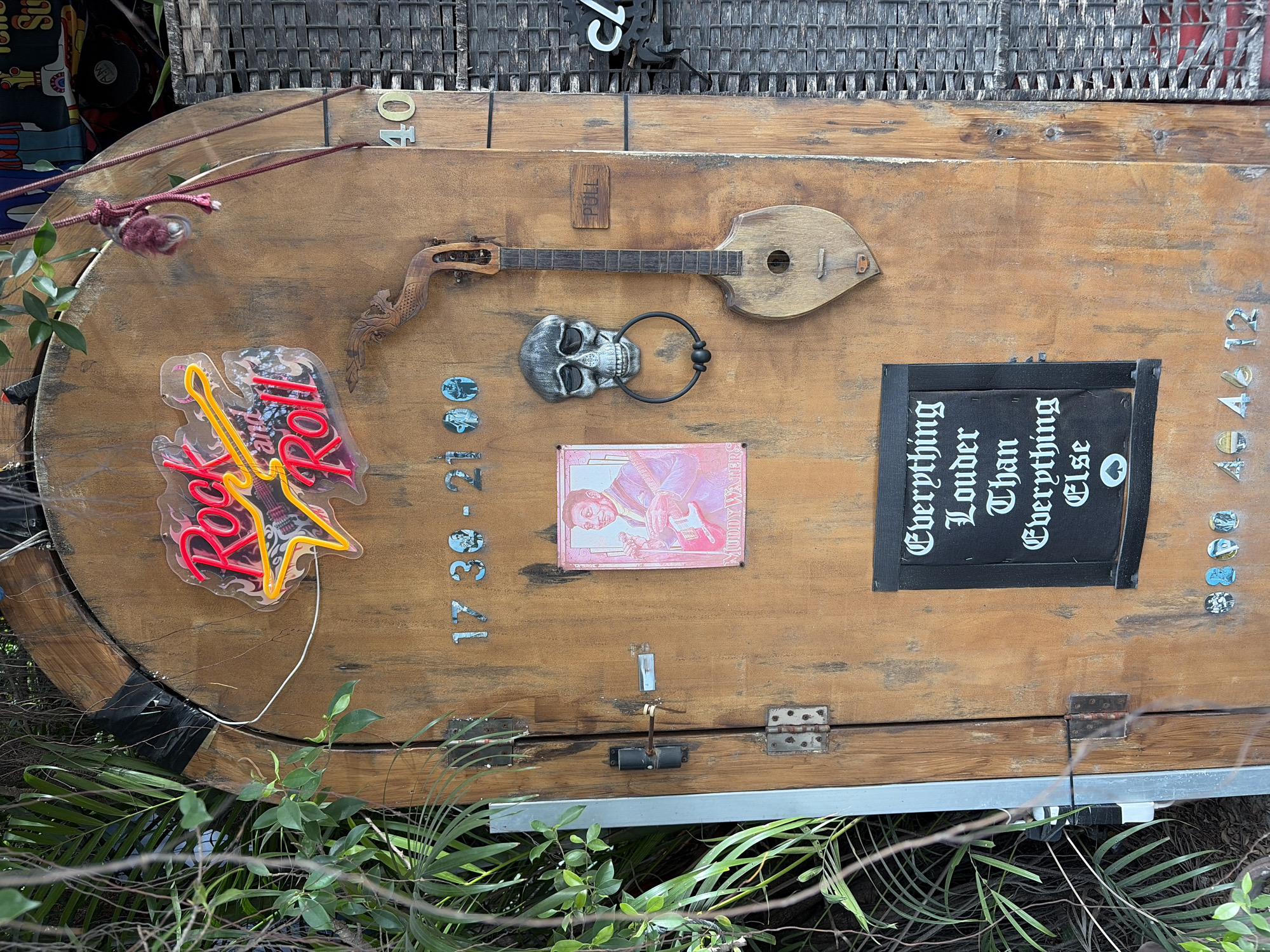 The image shows a weathered wooden door with several decorative elements. At the top, a neon sign reads "Rock and Roll" in red and yellow. Below the sign, a small, framed picture of a man playing a guitar is visible. To the right of the picture, a skull-shaped door knocker with a metal ring hangs from the door. A small, wooden guitar is mounted vertically on the door, to the right of the skull knocker. Above the guitar, a sign reads "PULL." Further to the right, the number "40" is affixed to the door. At the bottom of the door, a black sign with white gothic lettering states "Everything Louder Than Everything Else." Several small, circular objects, possibly coasters or buttons, are arranged near the bottom of the door. The door is set within a rustic environment, with greenery visible on the left side and a woven material on the right. The scene appears to be an exterior entrance, possibly to a establishment with a rock and roll theme. The location of Baan Bo Phut, Thailand, is not discernible from the visual elements of the image itself.