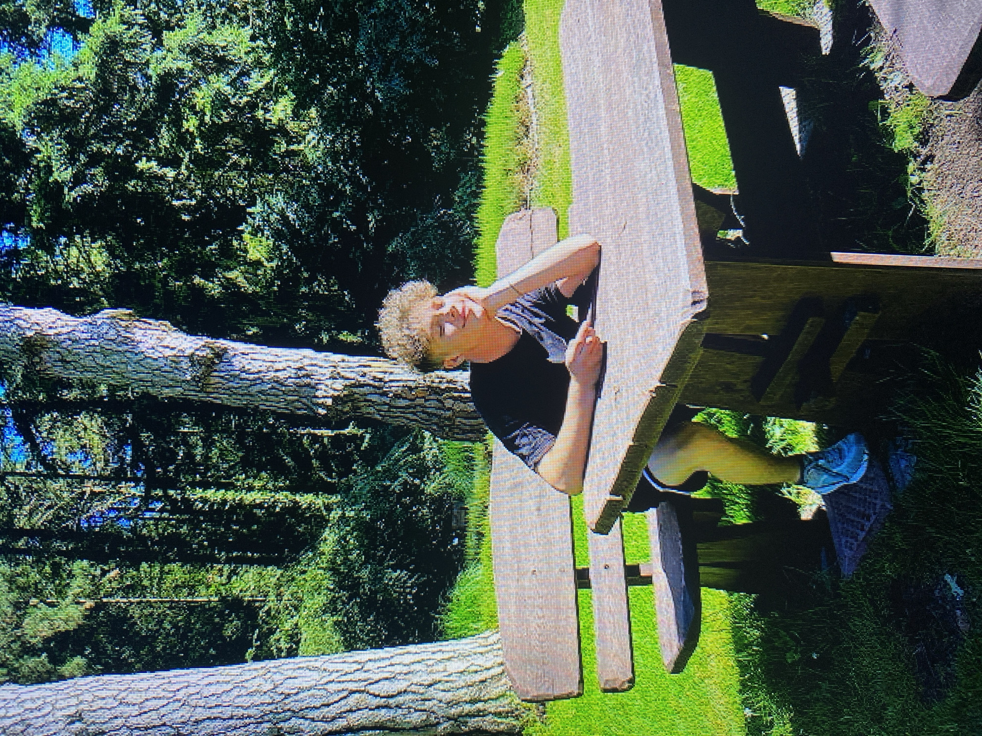 A young male with curly blonde hair, wearing a black t-shirt and grey athletic shorts with white stripes, sits at a wooden picnic table in a sun-dappled forest setting. He is leaning on his right hand, with his elbow on the table, and his head is tilted back slightly, with his eyes closed, suggesting a moment of rest or contemplation. His left hand rests on the table. He is wearing white and grey athletic shoes. The picnic table and bench are made of dark wood and appear weathered. The background consists of tall trees with rough bark and dense green foliage. The ground is covered with bright green grass and patches of dry earth. The scene is illuminated by direct sunlight, creating strong shadows. The location is identified as being in the city of Poznan, Poland, likely in a park or wooded area within the city limits.