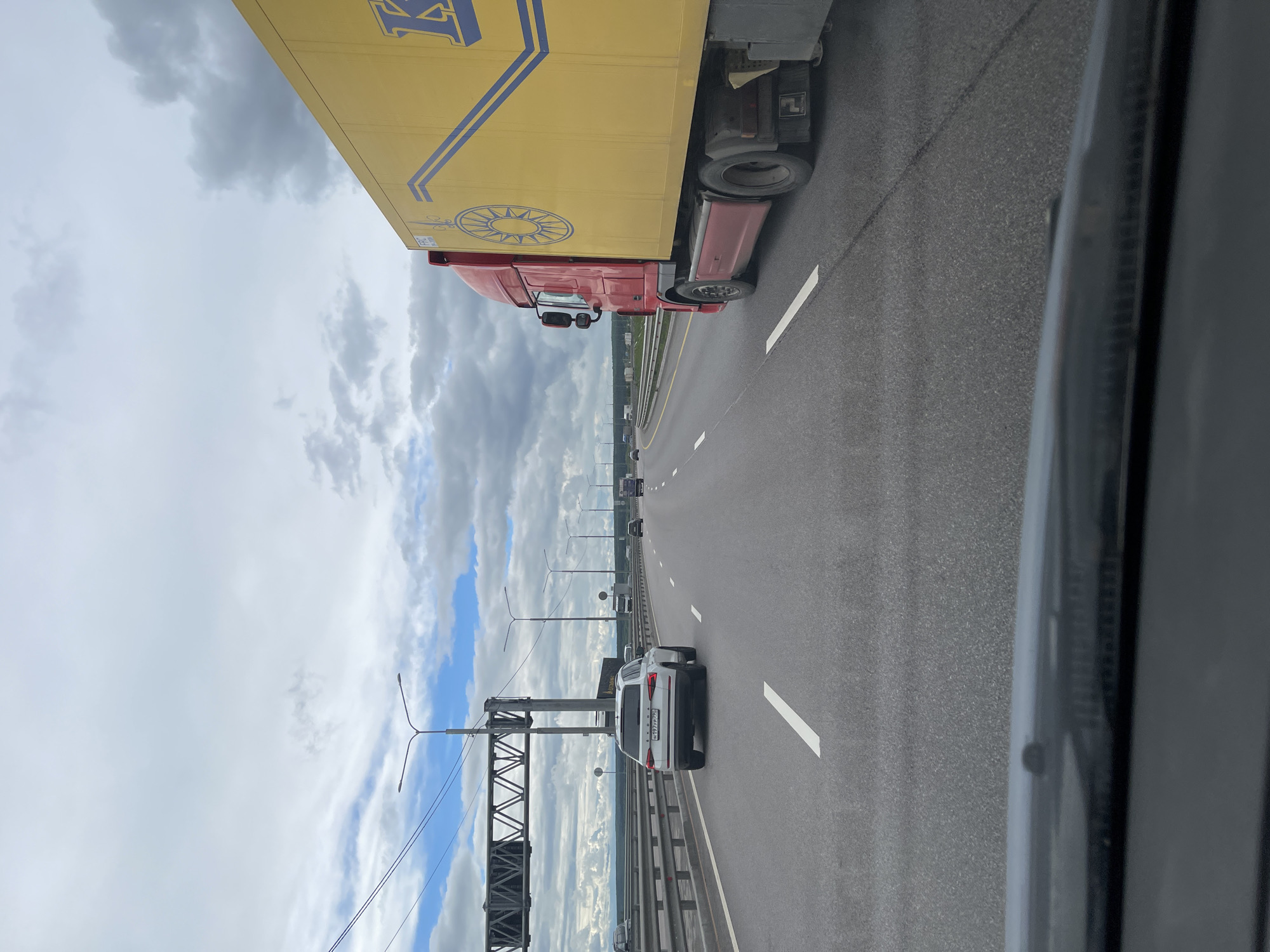 The image depicts a multi-lane highway in Taraskovo, Russia, with a large yellow semi-truck occupying the right lane. A white car is positioned in the adjacent lane, slightly ahead of the camera's perspective. Additional vehicles are visible further down the road in the distance. Overhead, a cloudy sky with patches of blue is visible, with roadside infrastructure like lampposts and bridge supports lining the highway. The image appears to be taken from inside a vehicle, with part of the car's interior visible in the foreground.