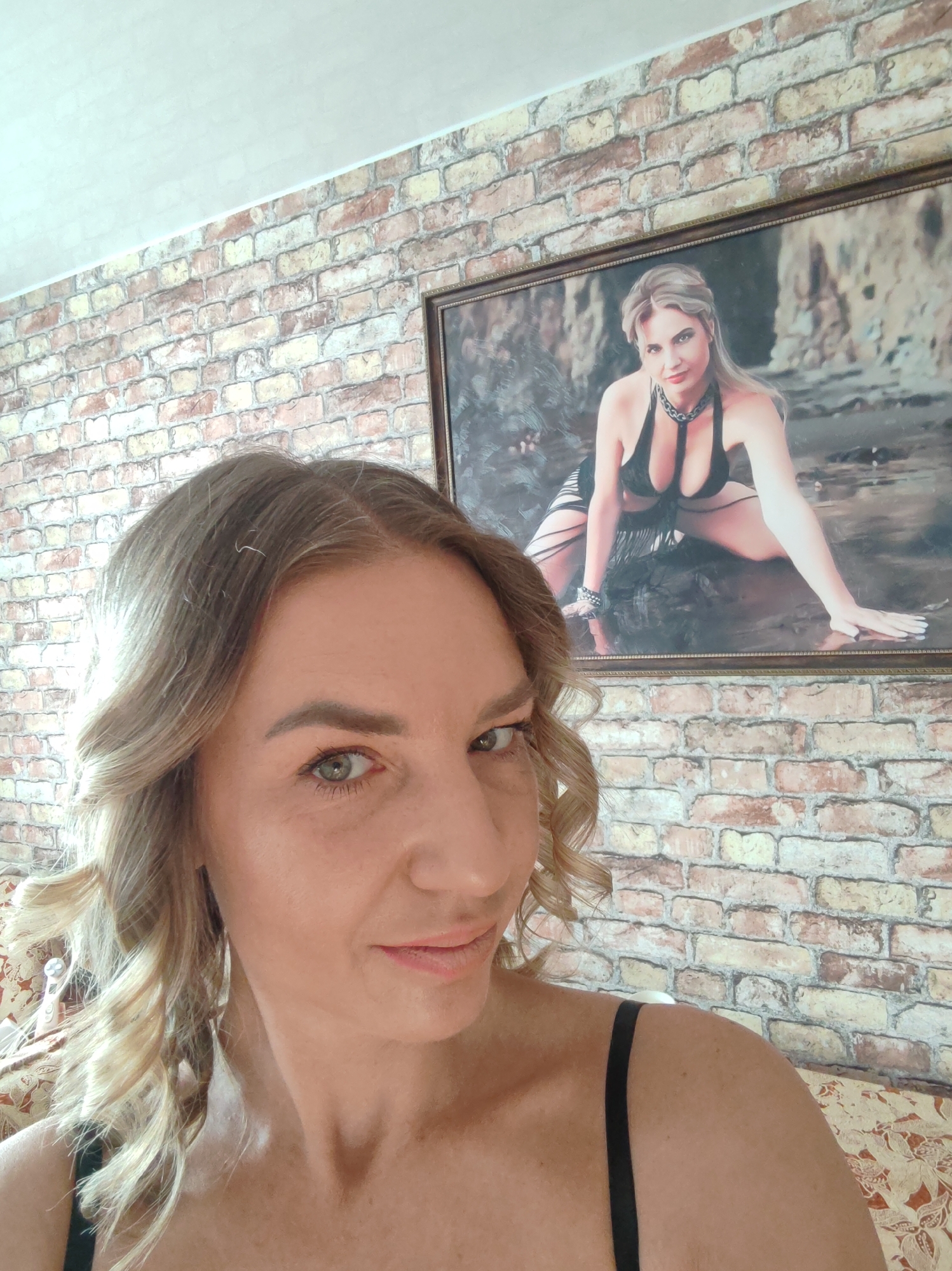 A woman is taking a selfie indoors. She is fair-skinned with light blonde hair and hazel eyes. She is wearing a black top. In the background is a brick-patterned wall. A framed picture hangs on the wall behind her, depicting a woman in a black bikini top and black accessories. It's impossible to determine the exact location in Petropavlovsk-Kamchatsky, Russia, without further contextual information.