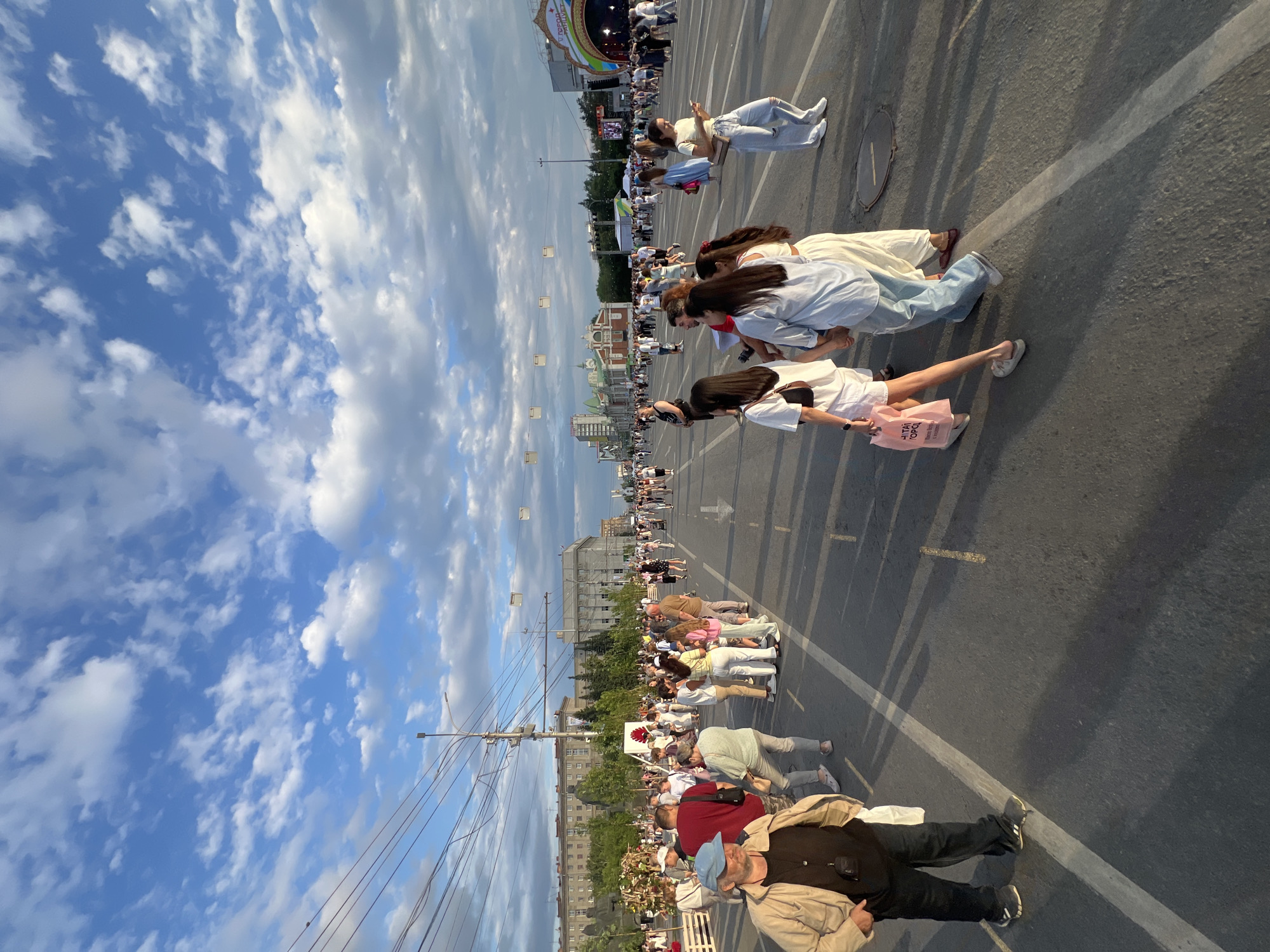 The image captures a wide, paved street in Novosibirsk, Russia, filled with a diverse crowd of people walking and congregating. The street appears to be temporarily closed to traffic, serving as a pedestrian thoroughfare. On the left side of the image, tall, imposing buildings line the street, some of which appear to be older, classic architectural styles, while others are more modern. Trees are planted along the buildings, adding a touch of greenery to the urban landscape. Overhead, a complex network of power lines stretches across the sky, indicating the presence of electrical infrastructure. The sky is a vibrant blue, dotted with fluffy white clouds, suggesting fair weather.
In the foreground, a man wearing a beige jacket and a blue cap is walking towards the camera, with his hands in his pockets. Behind him, a group of people are walking in various directions, some in small clusters, others individually. A woman in a white outfit is visible carrying a pink shopping bag. Further down the street, more people are spread out, suggesting a lively event or a busy public space. In the distance, the scene continues with more buildings and a stage setup with a large, illuminated archway, hinting at a potential festival, concert, or public celebration. The overall atmosphere appears to be one of a warm, sunny day, with a significant number of people enjoying an outdoor gathering in the heart of the city.