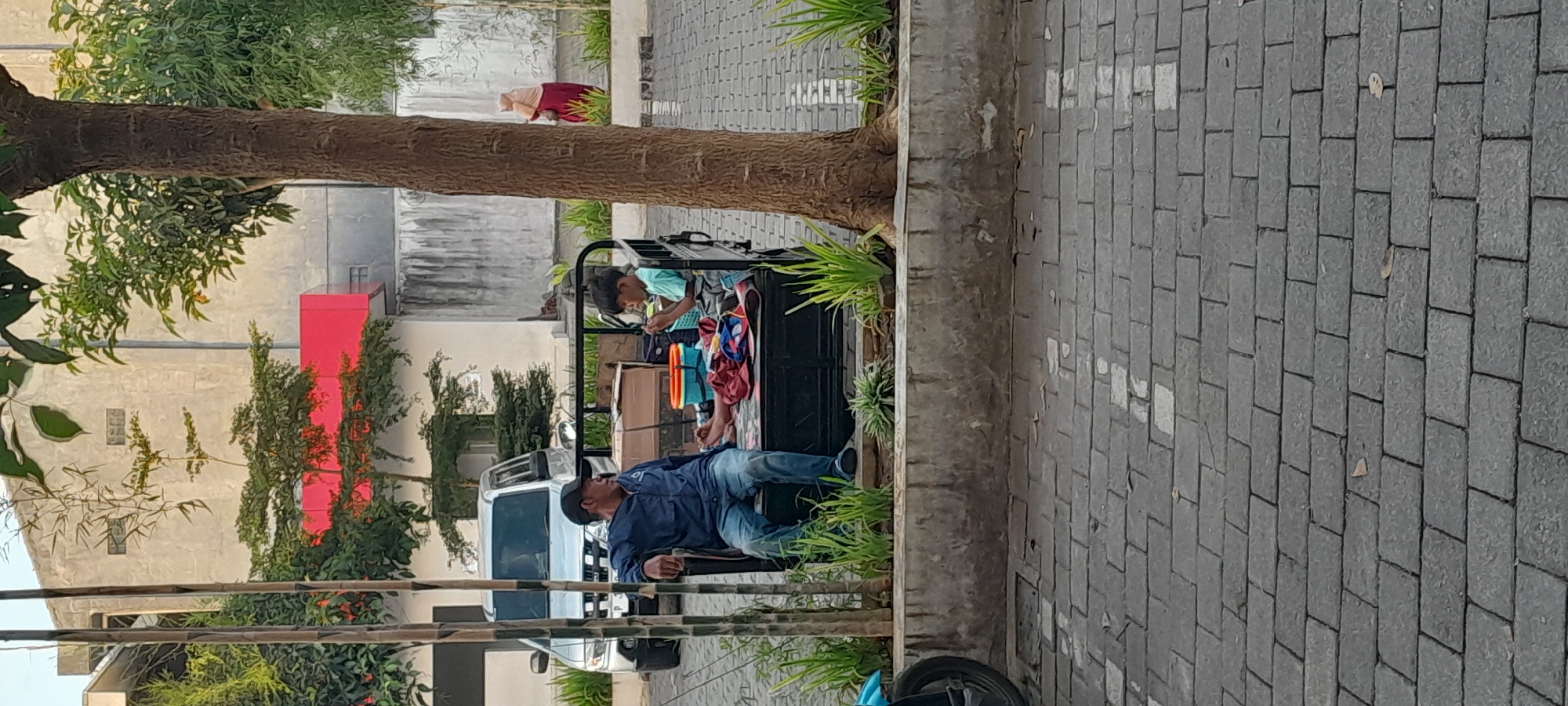 In Rogojampi, Indonesia, two individuals are seated in the back of a motorized vehicle. A man wearing a blue jacket and a black cap is seated on the left, with his legs extended forward. To his right, a child in a light blue t-shirt is seated, looking down at something in their hands. Various items, including bags and containers, are present in the vehicle. The vehicle is parked on a brick-paved surface. In the background, a silver car is visible, along with buildings and trees. A woman wearing a headscarf and a red garment is walking in the distance. The scene appears to be outdoors, possibly in a public area or a parking lot.