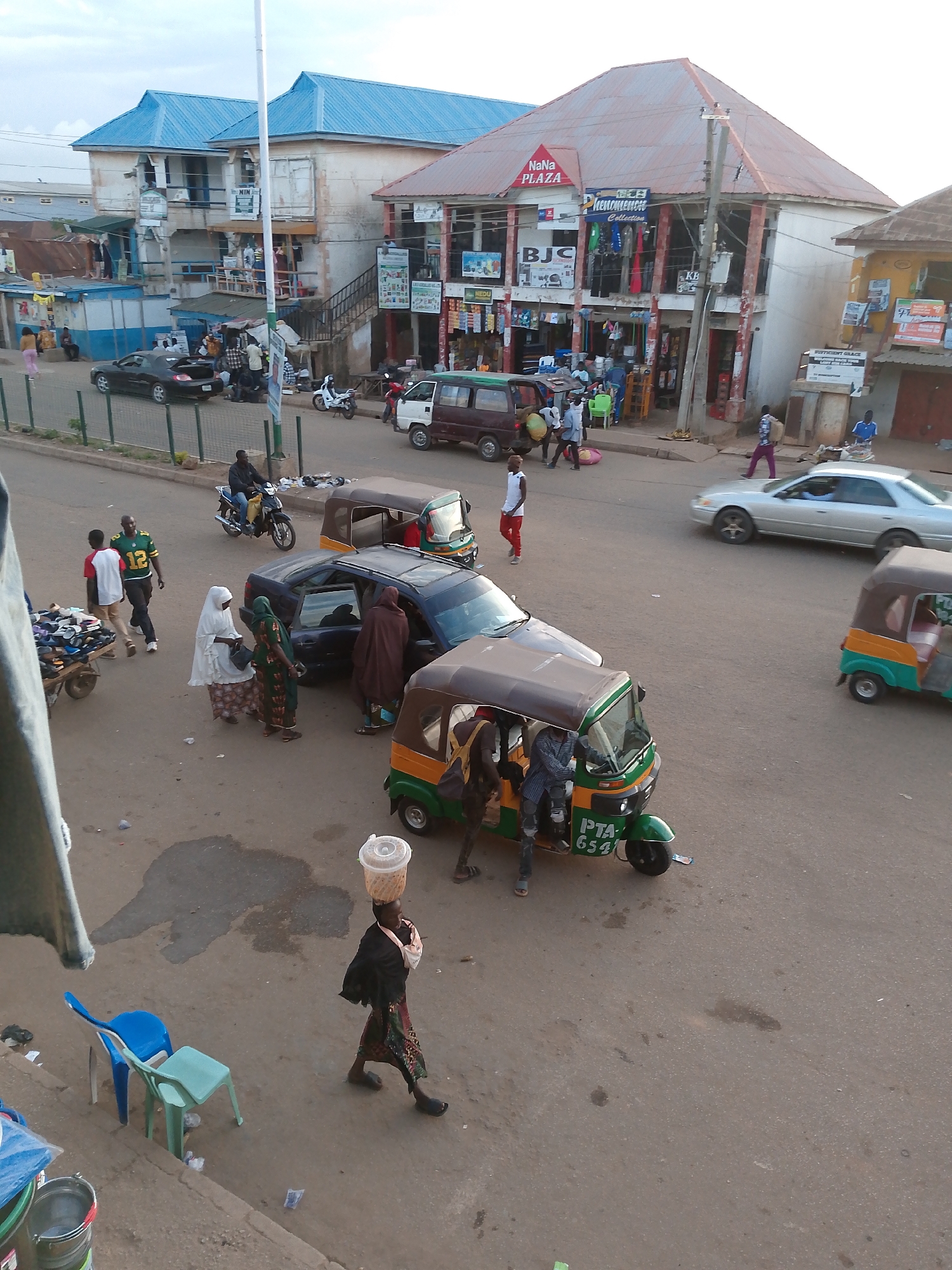 The image depicts a street scene in Bukuru, Nigeria, likely a market or commercial area. Several vehicles are present, including a dark-colored car, a white van, a green and yellow tuk-tuk, and another tuk-tuk partially visible on the right. Pedestrians are also visible, including a woman carrying a basket on her head and a man riding a motorcycle. Buildings line the street, some with storefronts displaying signs. The overall atmosphere appears busy and functional, with people engaging in daily activities. The presence of the tuk-tuks suggests a common mode of transportation in the area. The architecture of the buildings appears varied, with some featuring corrugated iron roofs and others having more modern designs. The ground surface is asphalt, indicating a paved road.