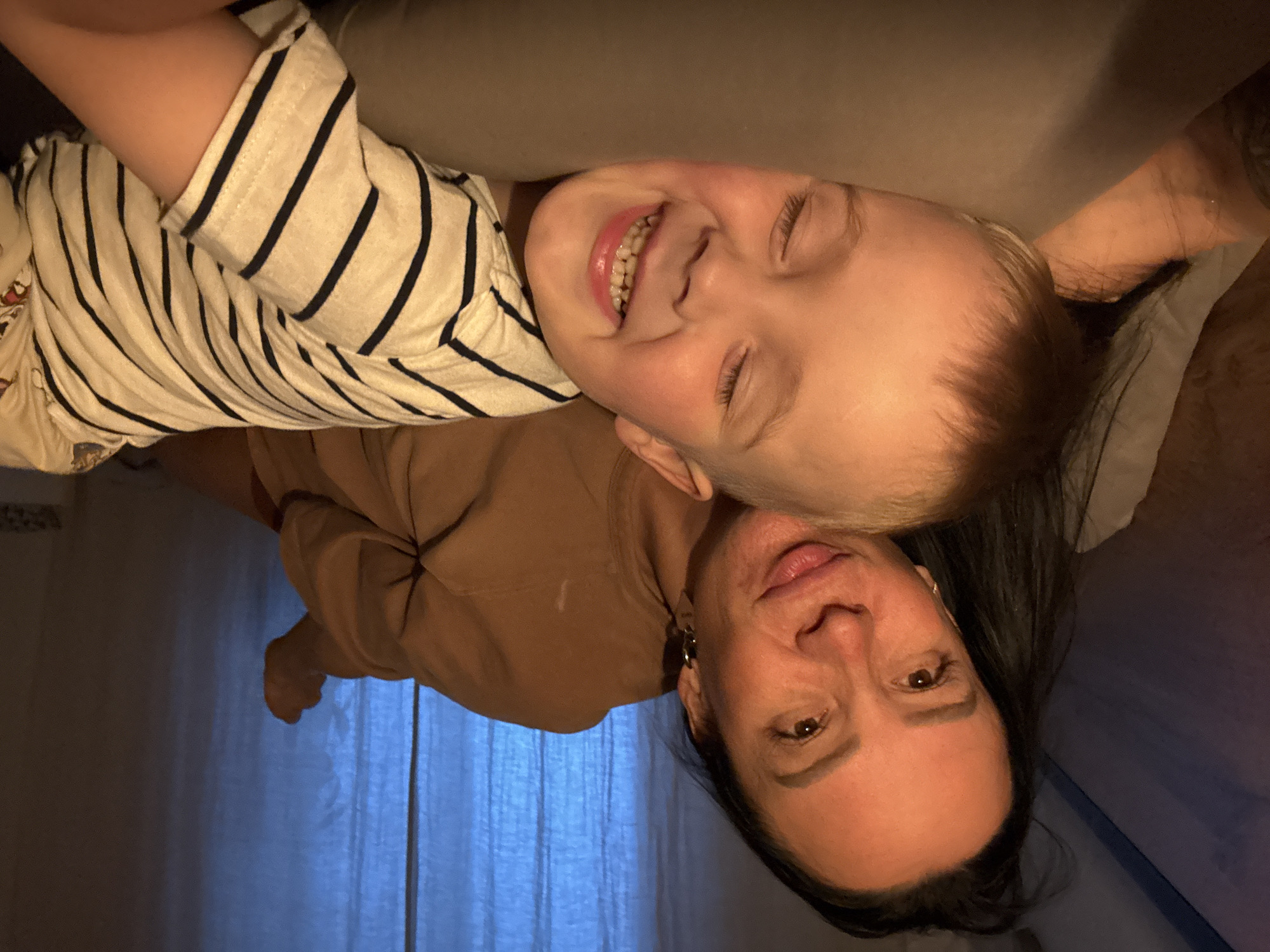 A woman and a child are lying down. The child is on top, smiling with closed eyes, wearing a white and black striped shirt. The woman is underneath, looking at the camera with dark hair and a brown shirt. Blue curtains are visible in the background. It is not possible to verify the location of the photo.