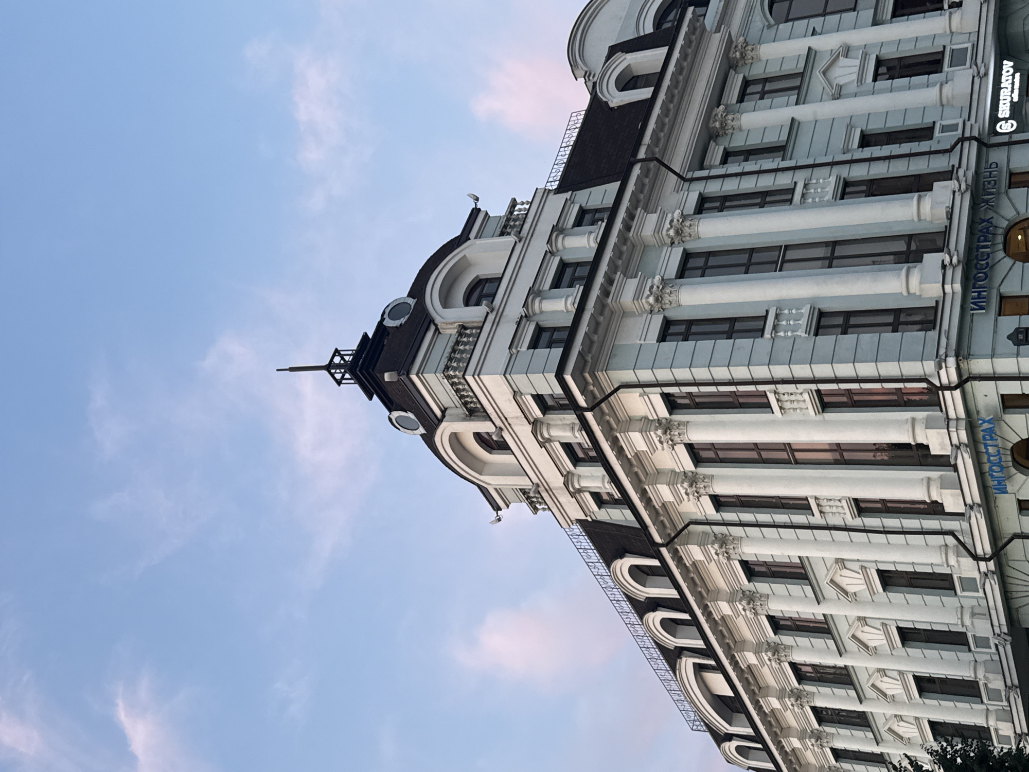 The image captures a close-up, low-angle view of the facade of the Ingostrakh building in Kazan, Russia, under a pale blue sky with wispy pink clouds. The building, a historical structure with classical architectural elements, features a light blue-grey exterior adorned with white columns and decorative moldings. The upper floors showcase arched windows and a dark mansard roof with dormer windows. At the very top, a dark spire with intricate metalwork rises towards the sky. Prominently displayed on the lower part of the building is a blue sign that reads "ИНГОССТРАХ ЖИЗНЬ," indicating the presence of the Ingostrakh insurance company. To the right of this sign, a smaller, illuminated sign with the logo "SKURATOV COFFEE" is visible, suggesting a coffee shop on the ground floor. No people are visible in the frame. The scene is illuminated by natural light, suggesting it was taken during daylight hours, possibly at dawn or dusk due to the soft hues in the sky.