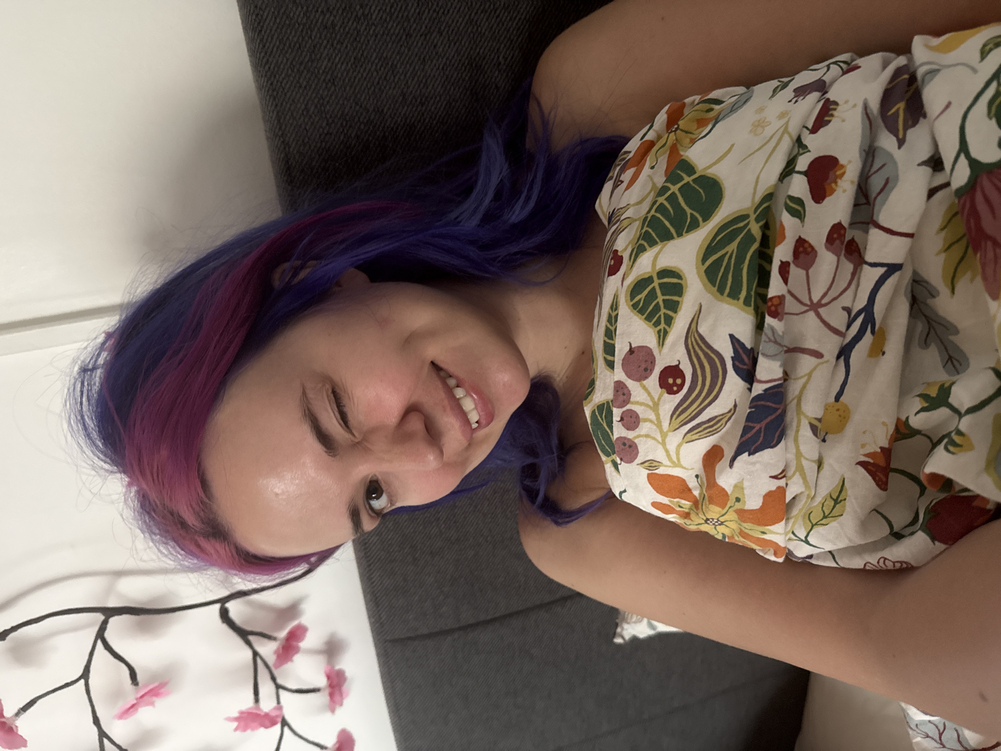 The image depicts a young woman with vibrant purple and pink dyed hair, smiling broadly with one eye playfully squinted. She is positioned against a dark gray upholstered surface, likely a sofa or bed. She is wearing a white top adorned with a colorful botanical pattern of leaves, berries, and flowers in various hues including green, red, orange, yellow, and blue. A decorative branch featuring pink artificial cherry blossoms is visible in the bottom-left of the frame, set against a white wall. The indoor scene is brightly and evenly lit, suggesting it is daytime. The woman's expression conveys happiness and playfulness. There is no visible text. The setting is likely a domestic interior in the City of Zagreb, Croatia.