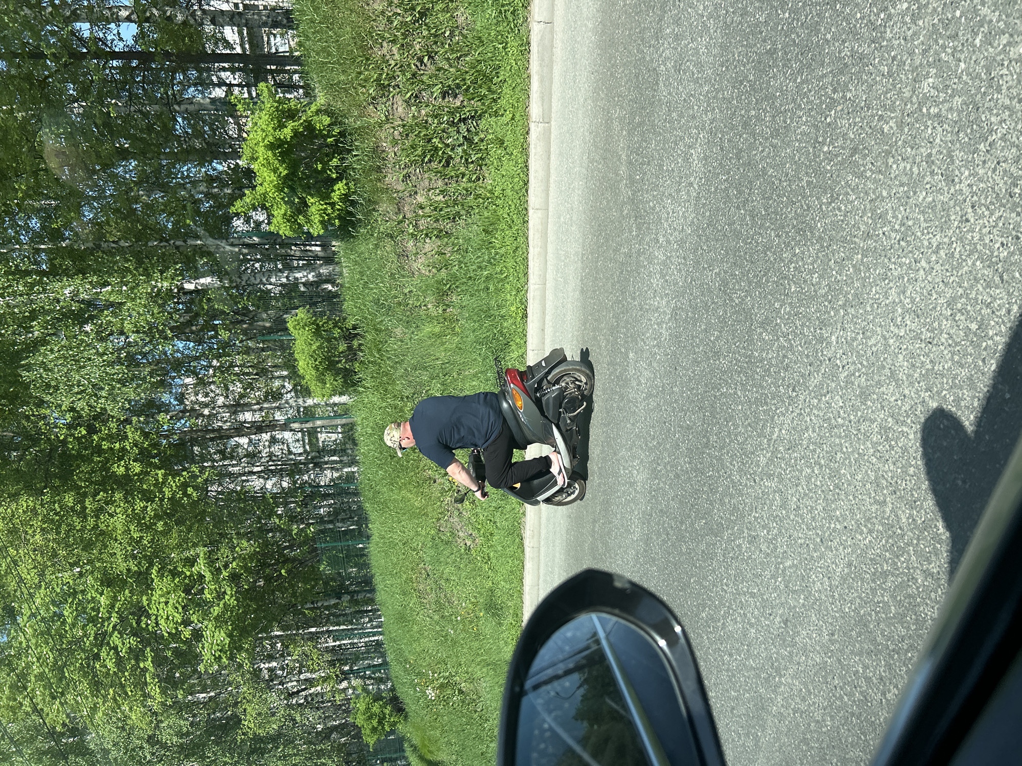 A man is riding a red and black scooter on a gray asphalt road in Izhevsk, Russia. The man is wearing a navy blue shirt, black pants, flip flops, and a camouflage hat. A green grassy embankment lined with birch trees is on the left side of the road. A car's side view mirror is visible in the lower right corner of the image, suggesting the photo was taken from inside a vehicle.