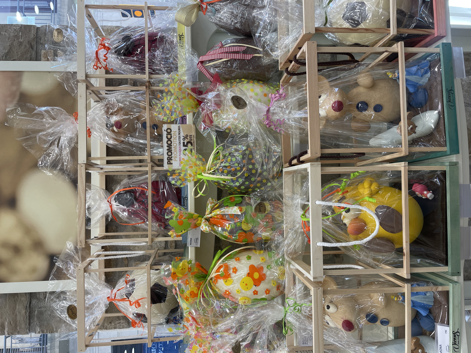 The image depicts a display of Easter confectionery in what appears to be a shop window. The confectionery includes chocolate Easter eggs wrapped in cellophane and decorated with ribbons, bows, and various designs. Some of the eggs feature designs of flowers, dots, and other patterns. Additionally, there are chocolate figures of animals and other characters encased in wooden crates, also wrapped in cellophane. The location may be Barcelona, Spain, given the Easter items and language on the "PROMOCIO" sign that reads "figures pasqua i easter figures". The confectionery is arranged in a multi-tiered display of wooden frames. Signs indicate pricing and sales promotions. The environment is a retail setting, likely a confectionery shop or a department store with a confectionery section.