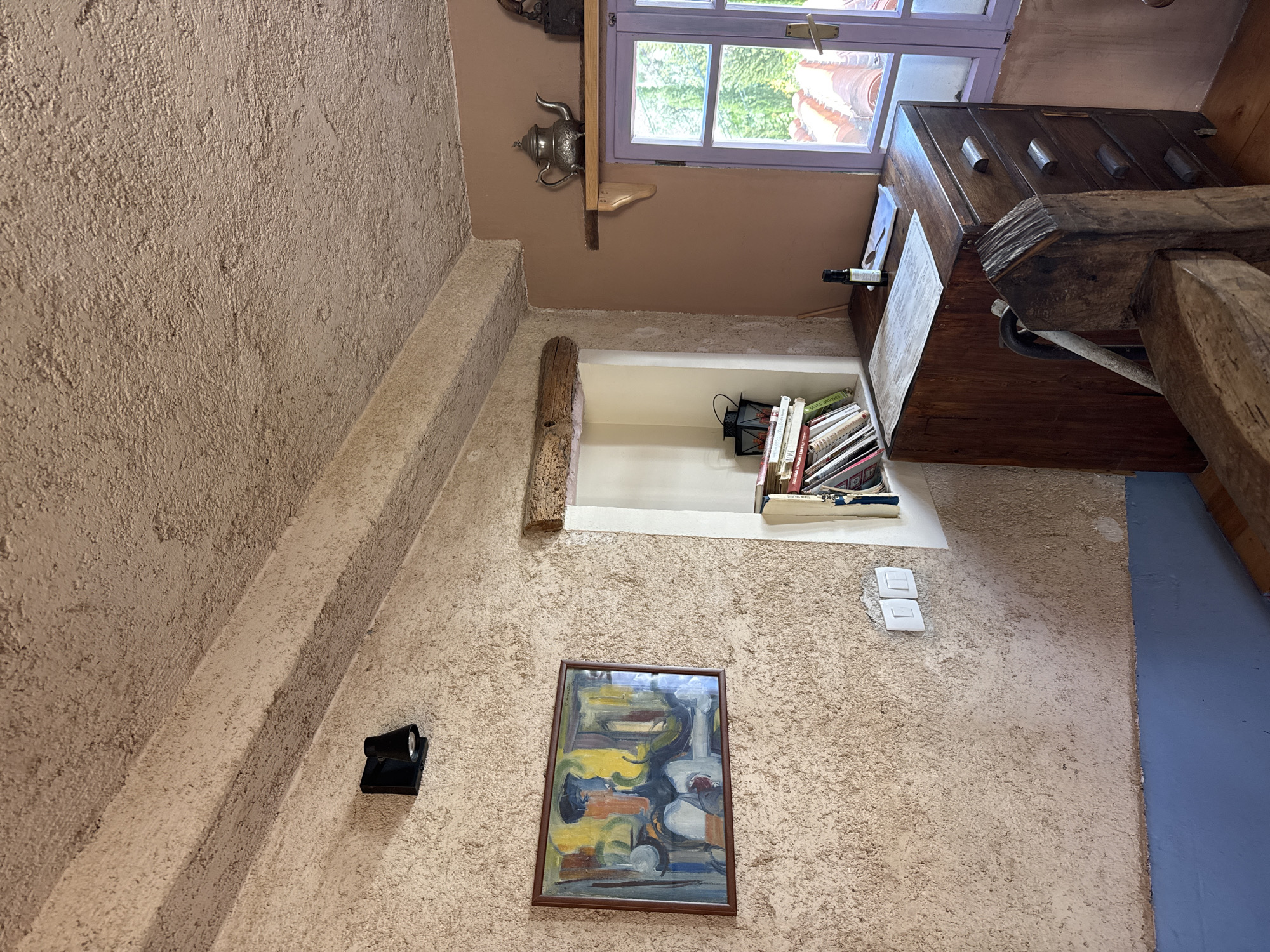 This image depicts an interior scene within a building, characterized by textured walls, likely stucco or a similar material. The color palette is predominantly earthy tones of beige and brown. The lighting appears to be a mix of natural light filtering in from a window and artificial light.
The main architectural features include a staircase leading upwards, a window with a lilac frame through which a blurred outdoor scene can be observed, and a built-in niche or shelf. In the niche is a collection of books and other objects.
Objects of interest are the silver teapot mounted near the window, a framed painting hung on the wall, an electrical spotlight fixture, and a pair of electrical switches. There is a piece of furniture, perhaps a chest of drawers, and a rough-hewn wooden beam in the corner.
The environment does not show any indication of where the image might be located, or any people present.