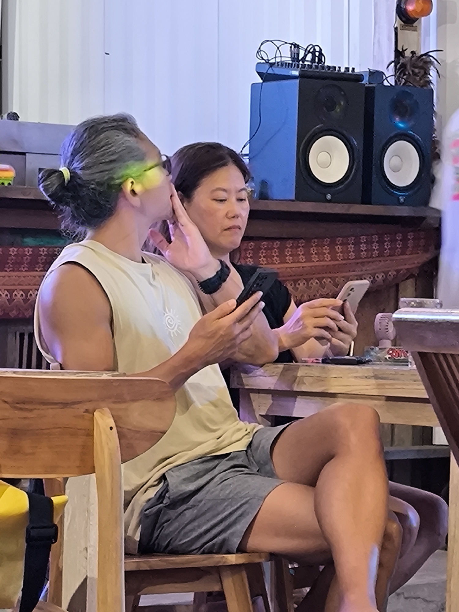 The image depicts two individuals seated at a wooden table. The person in the foreground is holding a smartphone and has their other hand raised to their face. They are wearing a tan tank top, grey shorts, and have grey hair pulled back in a ponytail. The other individual, sitting behind, is also holding a smartphone. They are wearing a black top. Behind them are two black speakers and a control panel. The background features a patterned tapestry. The location is in the city of Unknown, Unknown.