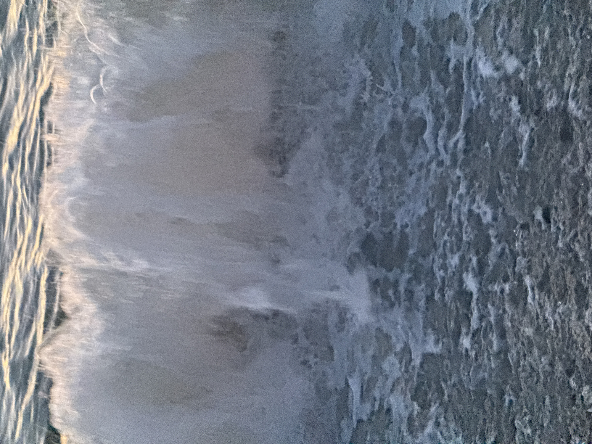 The image shows water, likely the sea, with a foamy wave breaking. The color is mostly gray with some white foam and a golden hue where the light reflects off the water's surface. The water appears turbulent and agitated. The location is presumed to be near Batumi, Georgia, given the prompt's instructions.